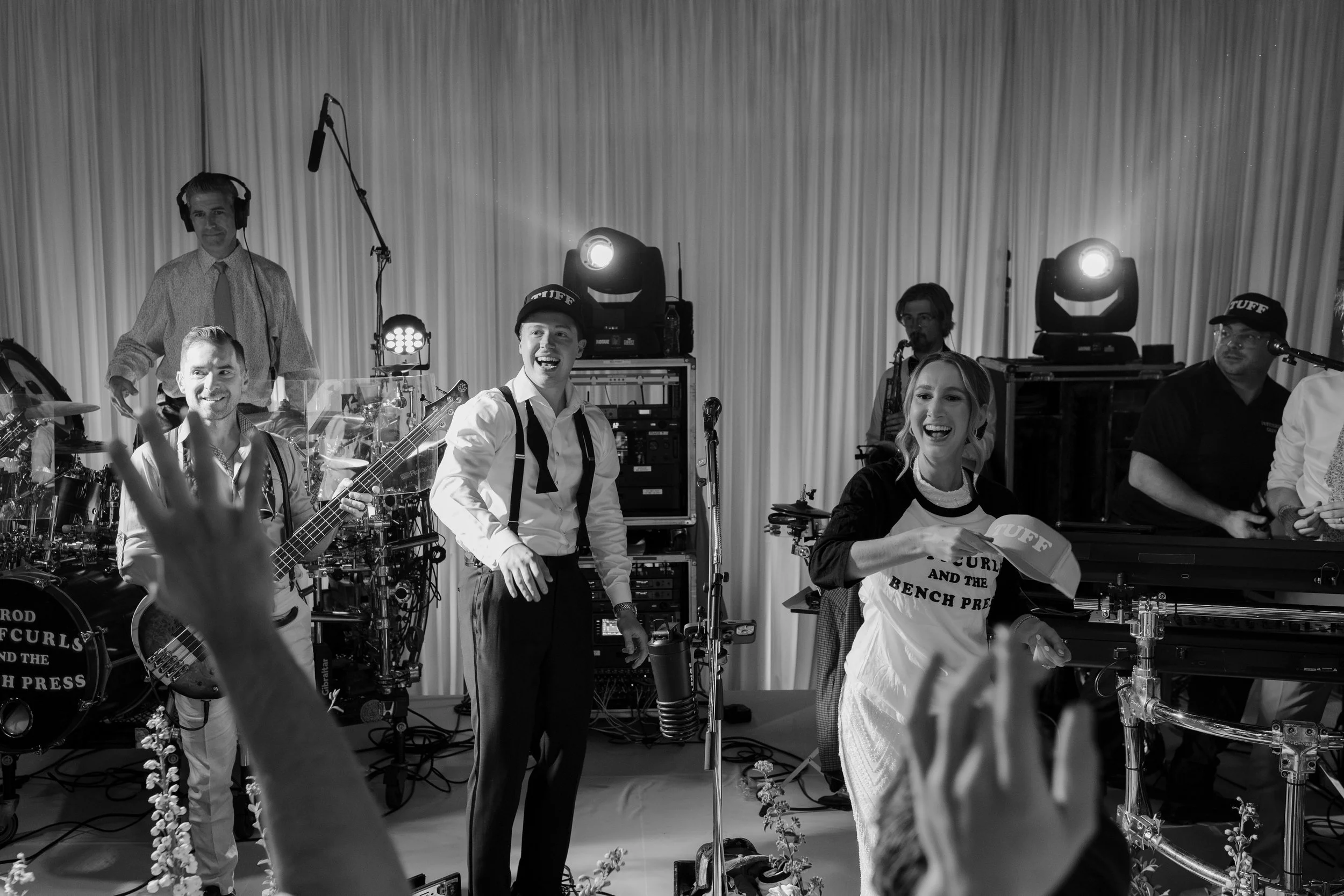 A black-and-white photo of a live music band performing on stage with smiling band members, musical instruments, and stage lights.