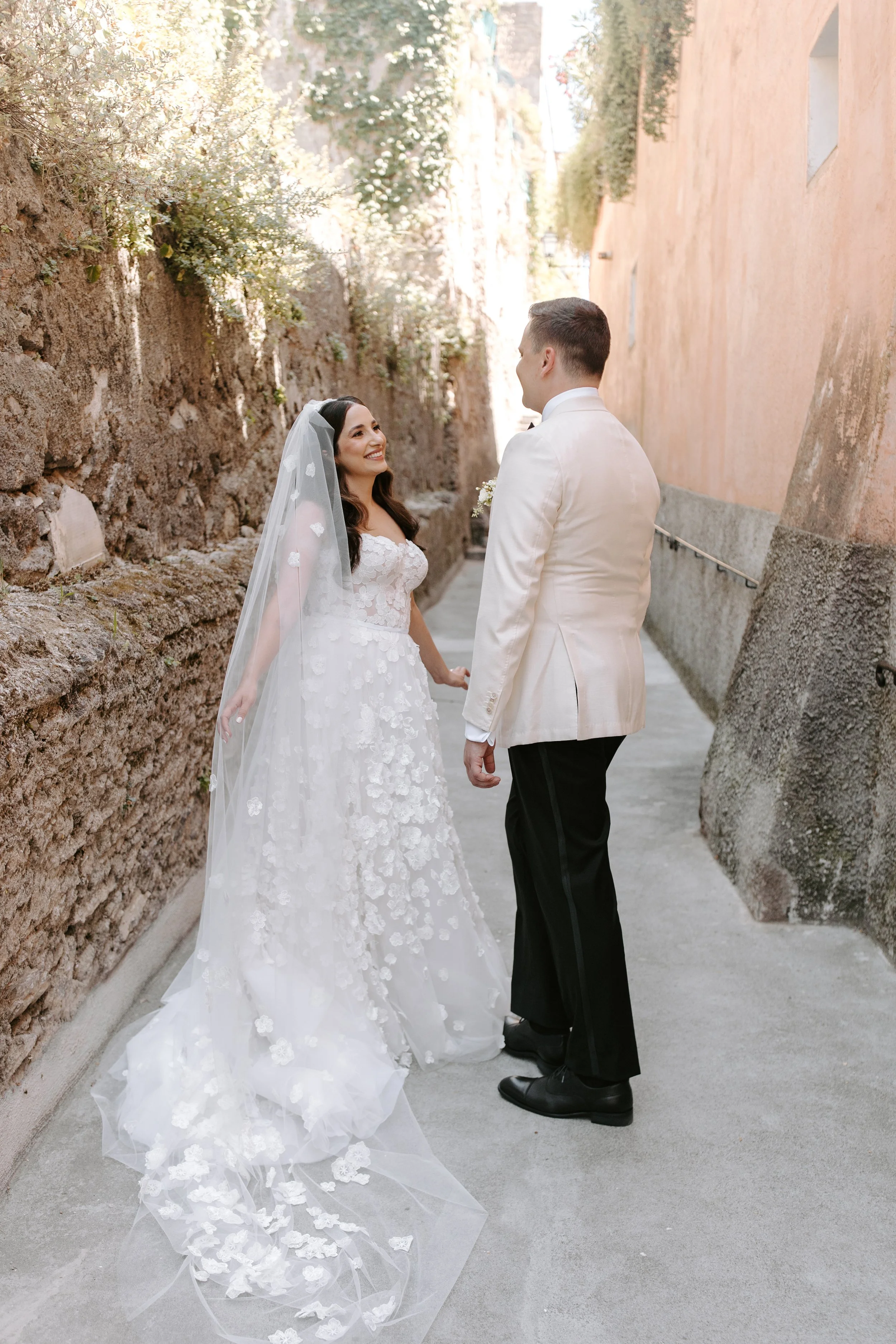 Bride and groom holding hands and smiling at each other outdoors, with a stone wall on one side and a building on the other.