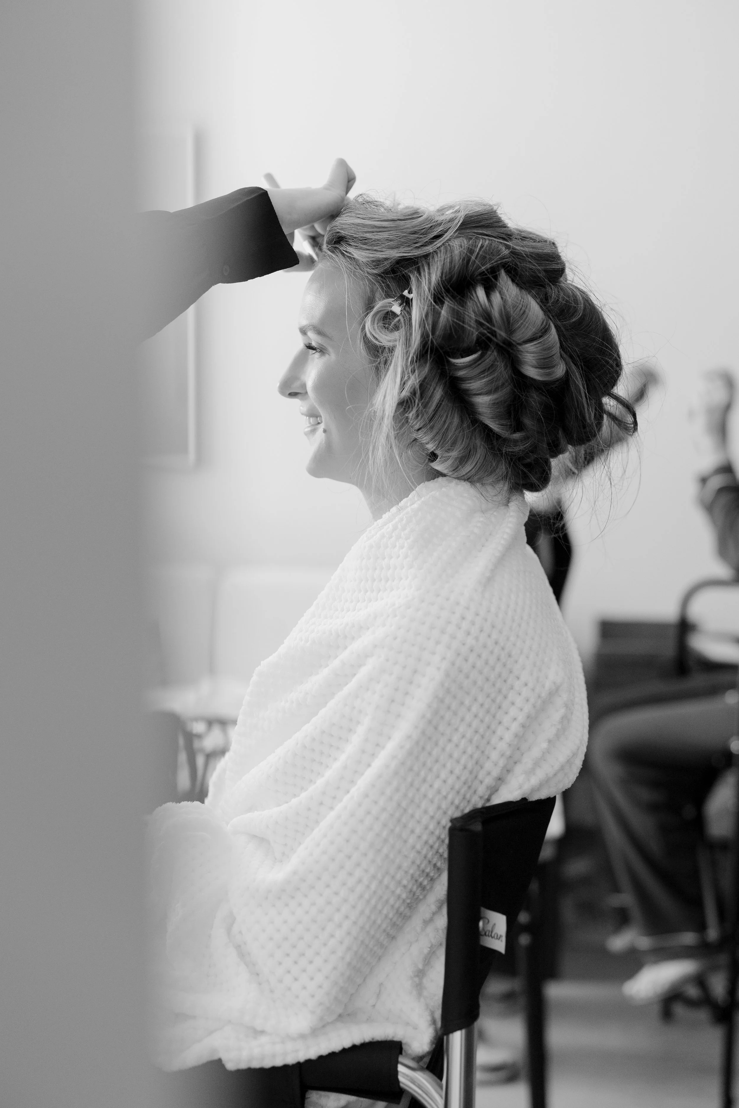 A woman with curled hair sitting in a chair, smiling, as someone applies hair rollers at a salon.