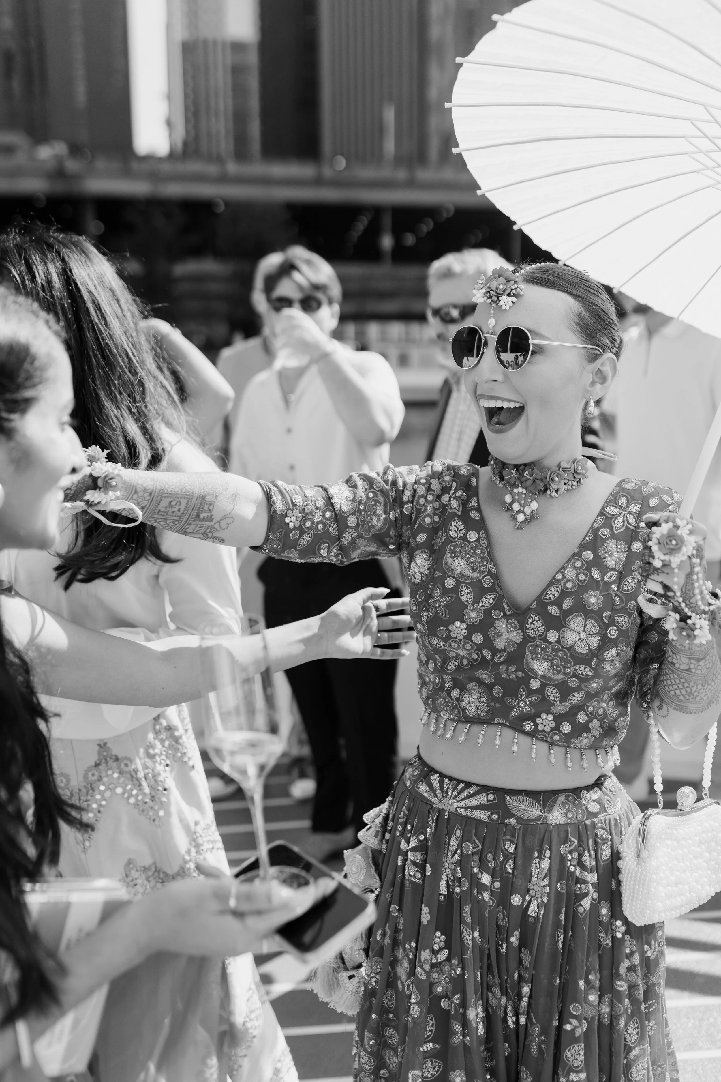 A woman wearing sunglasses, floral jewelry, and traditional attire is smiling and reaching out to greeting or shake hands with a person in a white dress, during an outdoor celebration under a paper parasol. Several other people are in the background.