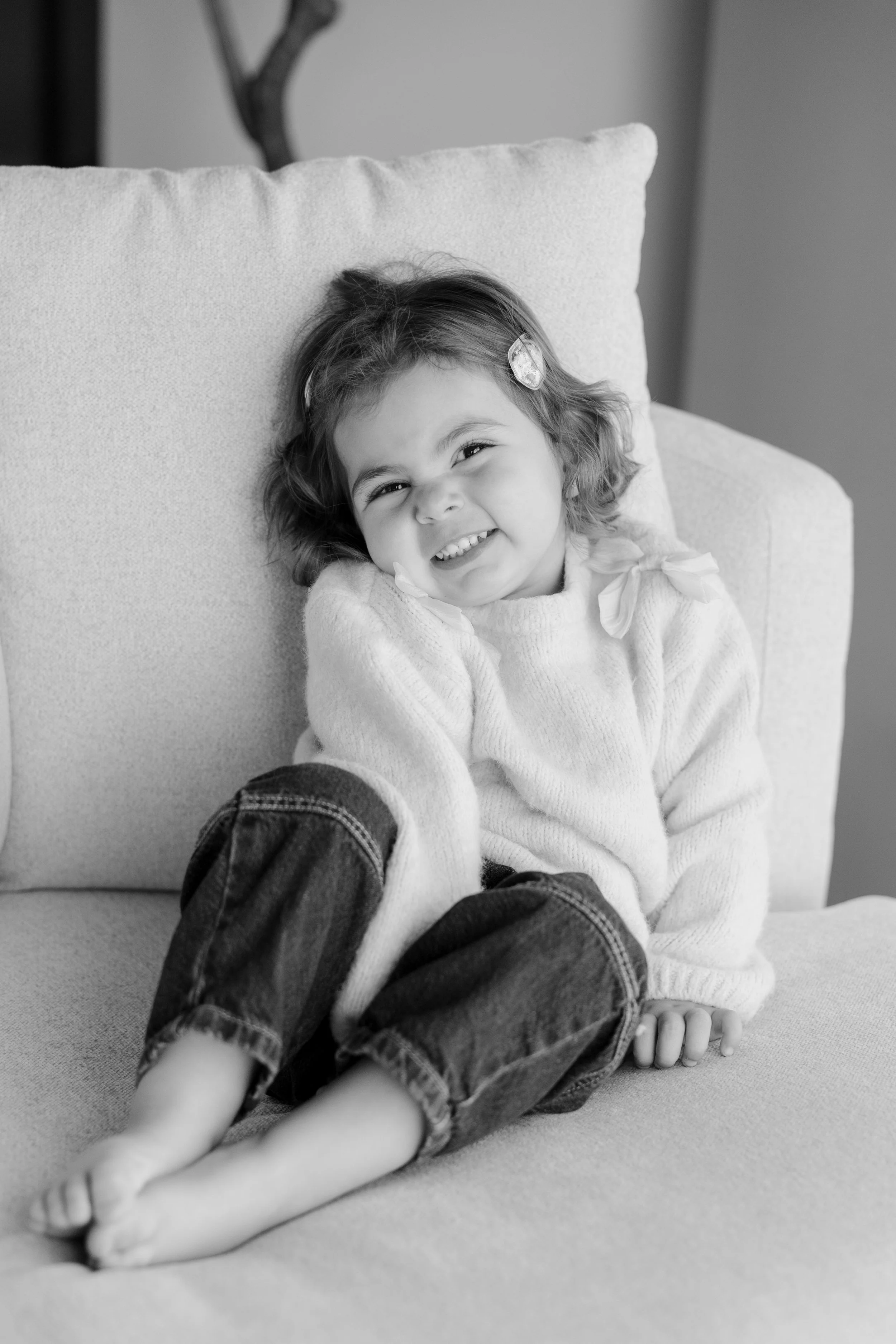 A young girl with curly hair and a hair clip, sitting on a couch, smiling at the camera in a black-and-white photo.