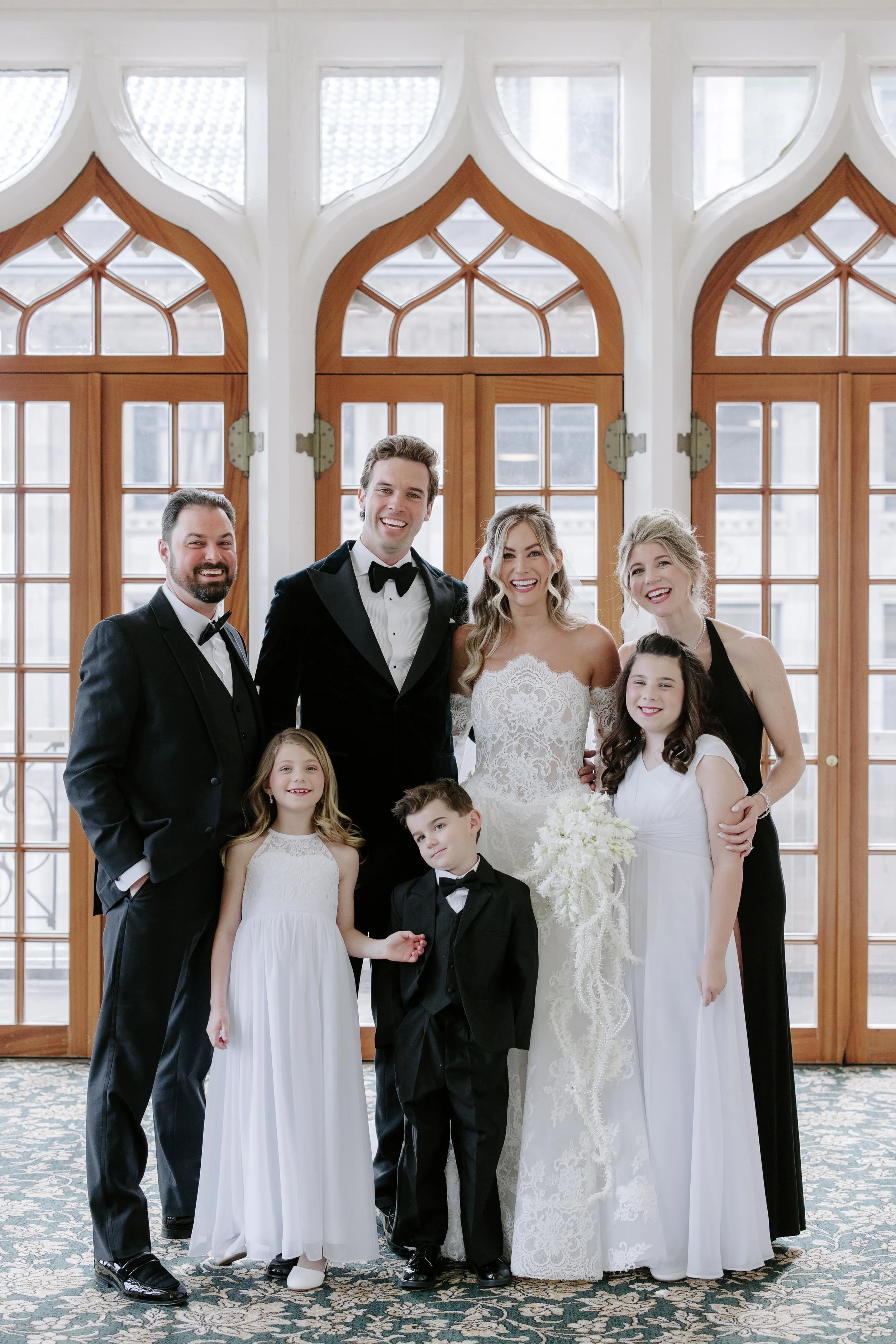 A group of people dressed for a wedding, including a bride in a white lace gown, a groom in a black tuxedo, and children in formal attire, standing in front of large wooden doors with decorative glass windows.