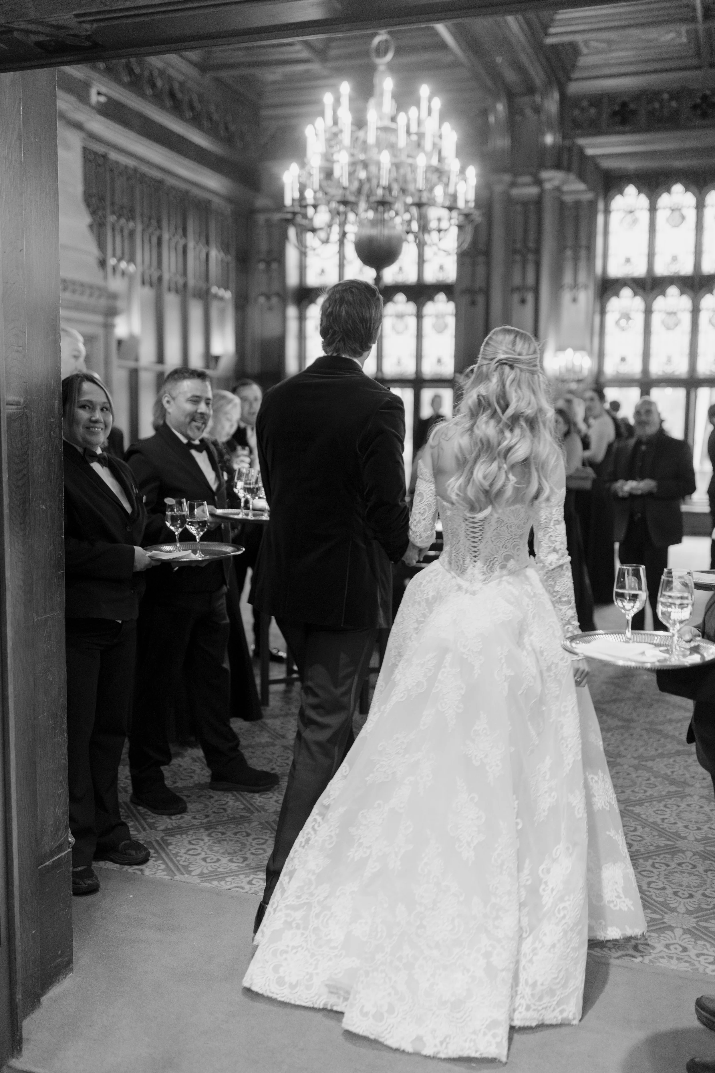 A bride and groom standing together at their wedding, with wedding guests in the background in a grand, wood-paneled room with large stained glass windows and a chandelier.