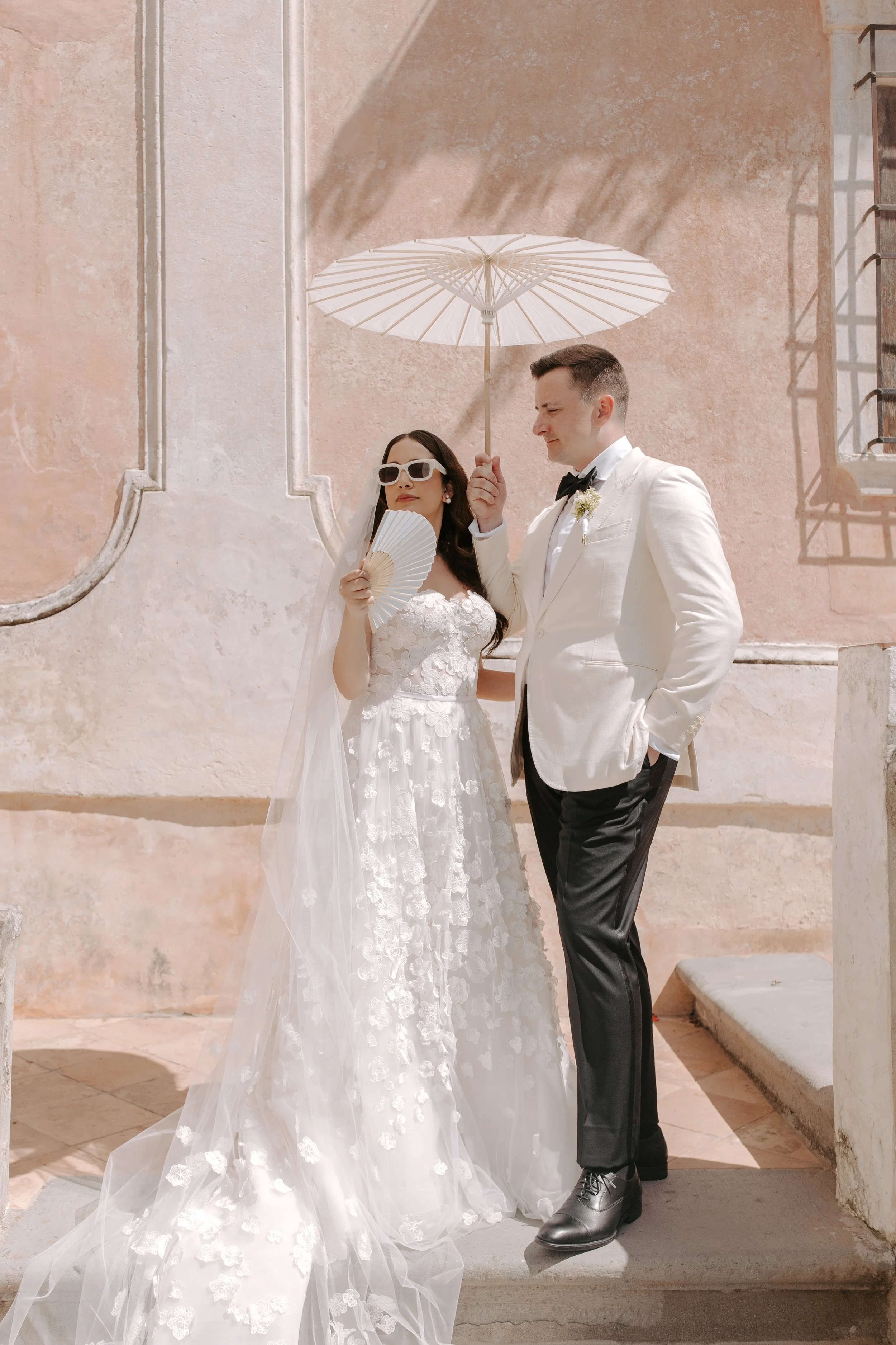 A bride and groom dressed in wedding attire outdoors, under a paper parasol, with a pink textured wall behind them.