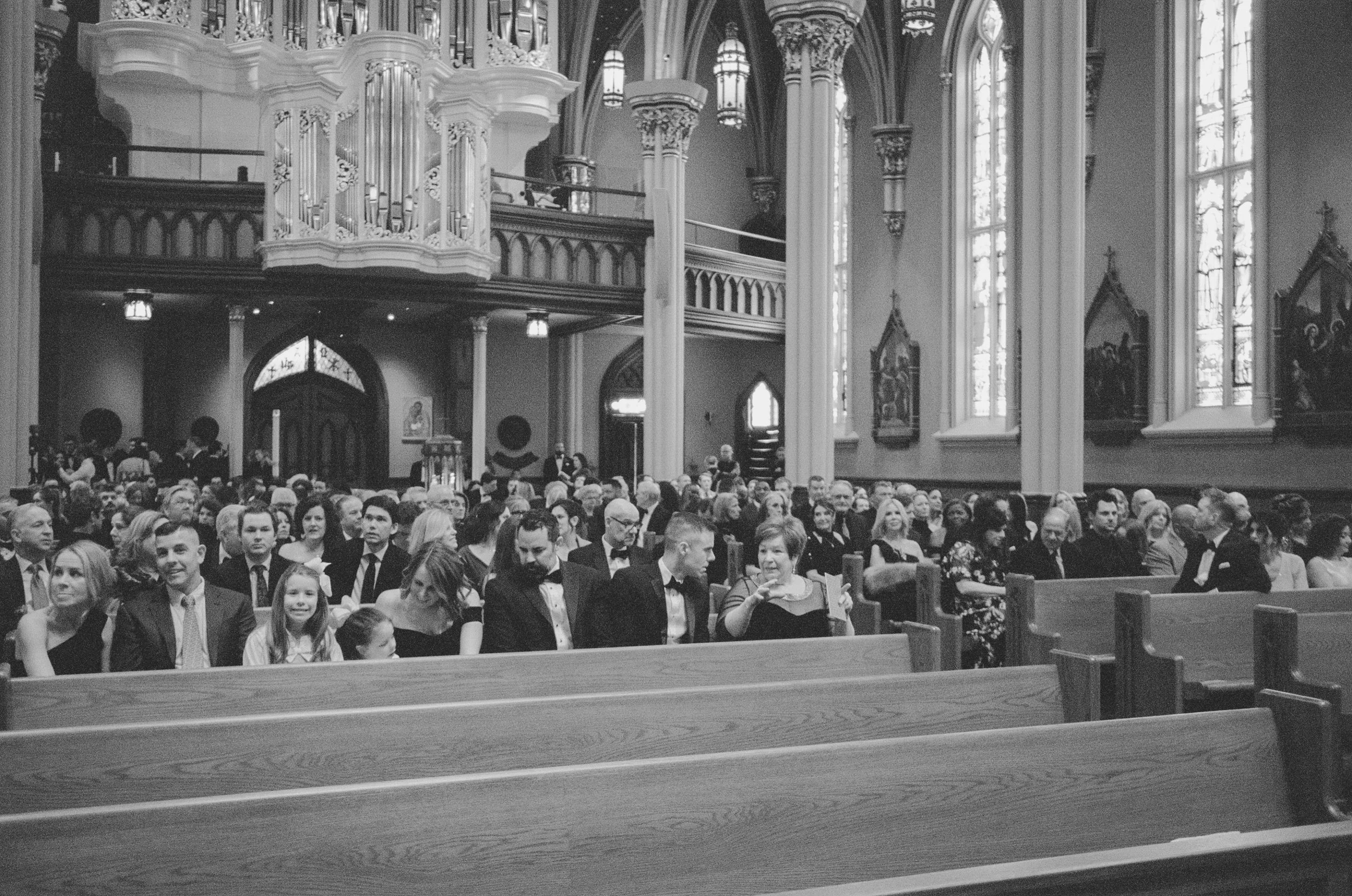 Inside a church filled with people seated in pews, attending a service or event. The church has tall, ornate columns, large stained glass windows, and an organ displayed in the background.