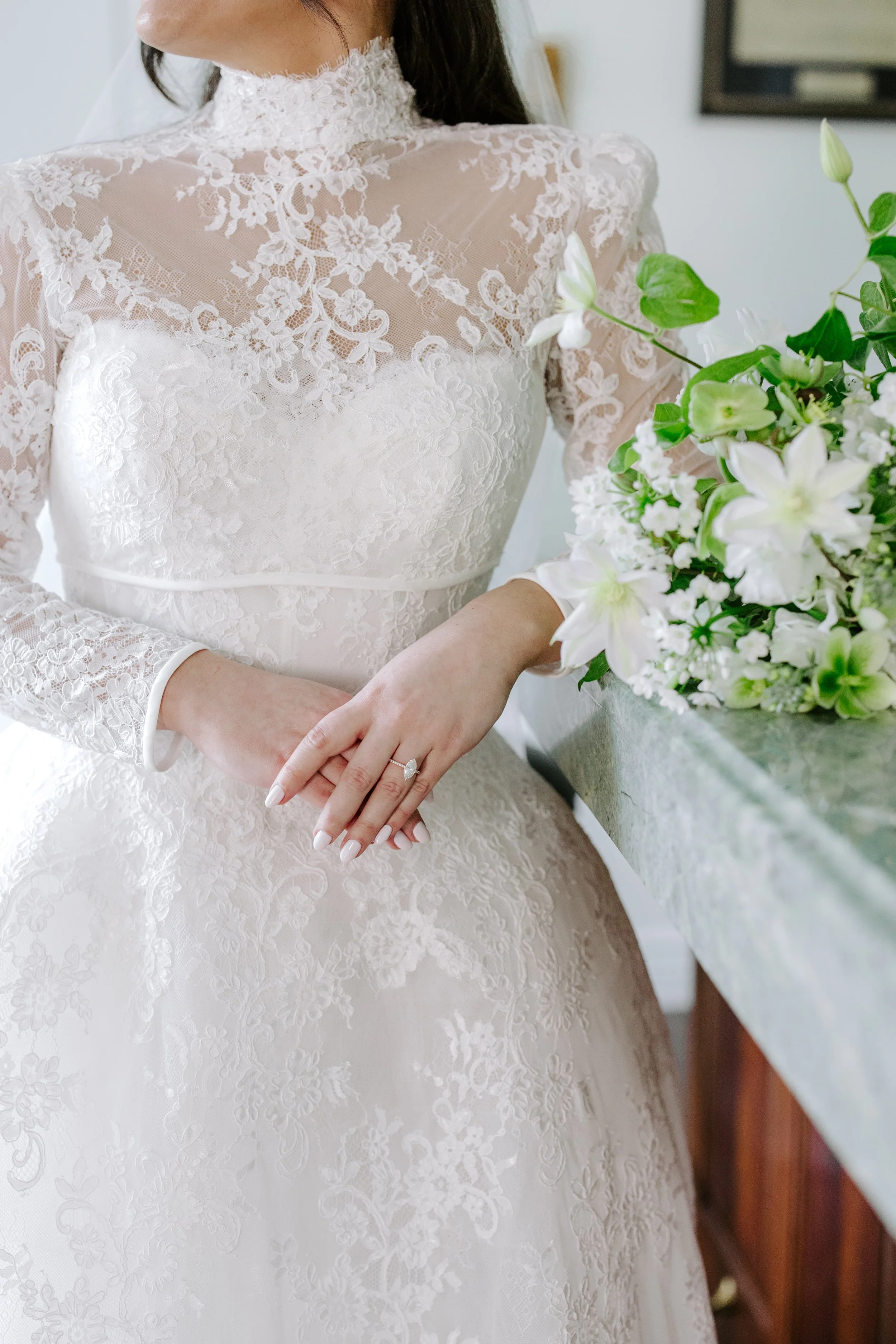A bride in a white lace wedding dress with long sleeves and a high lace neckline. She is holding her hands together with a ring on her finger, standing near a bouquet of white flowers with green leaves on a marble surface.