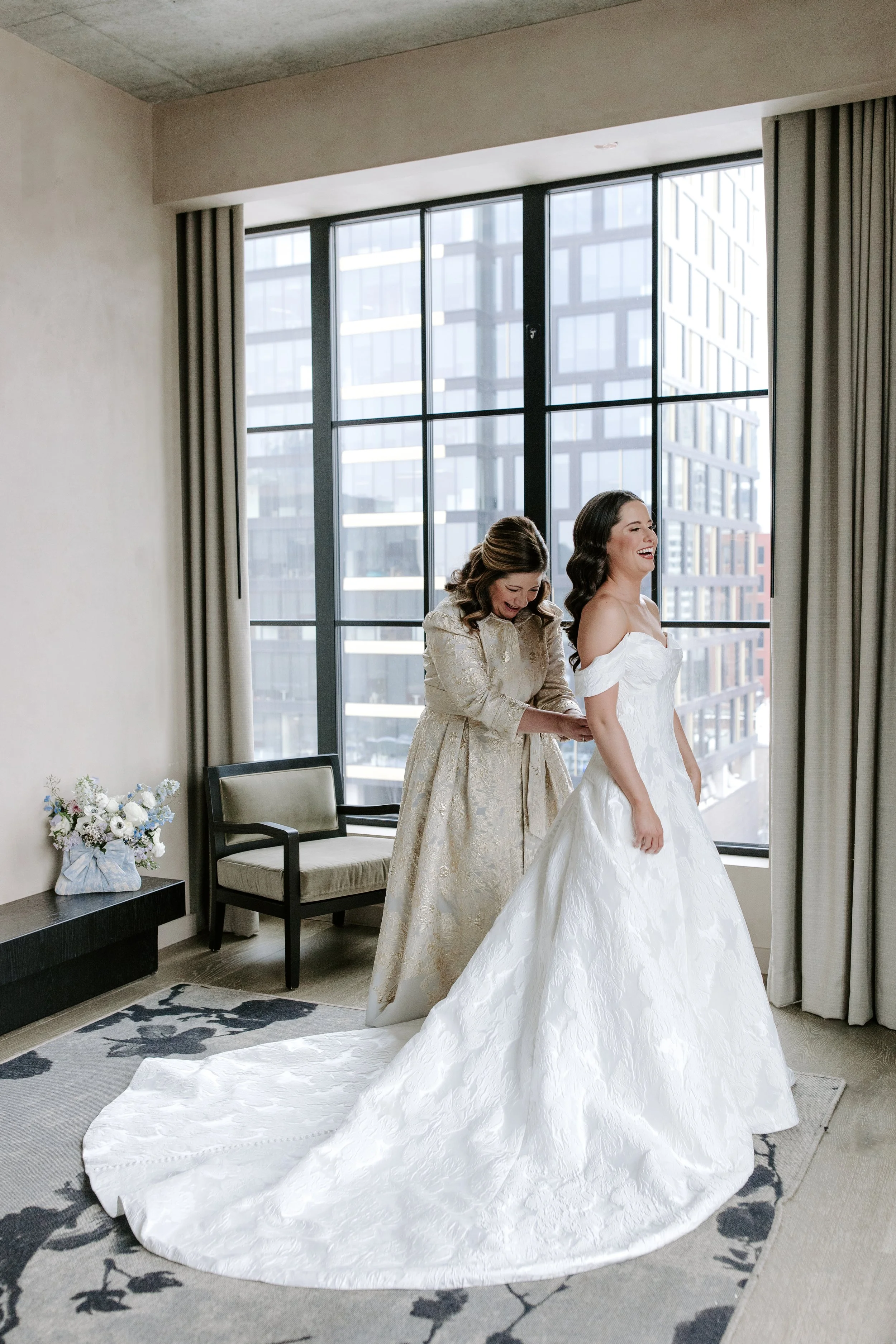 Two women, one in a wedding dress and the other in a gold gown, are laughing and smiling in front of a large window with city buildings in the background.