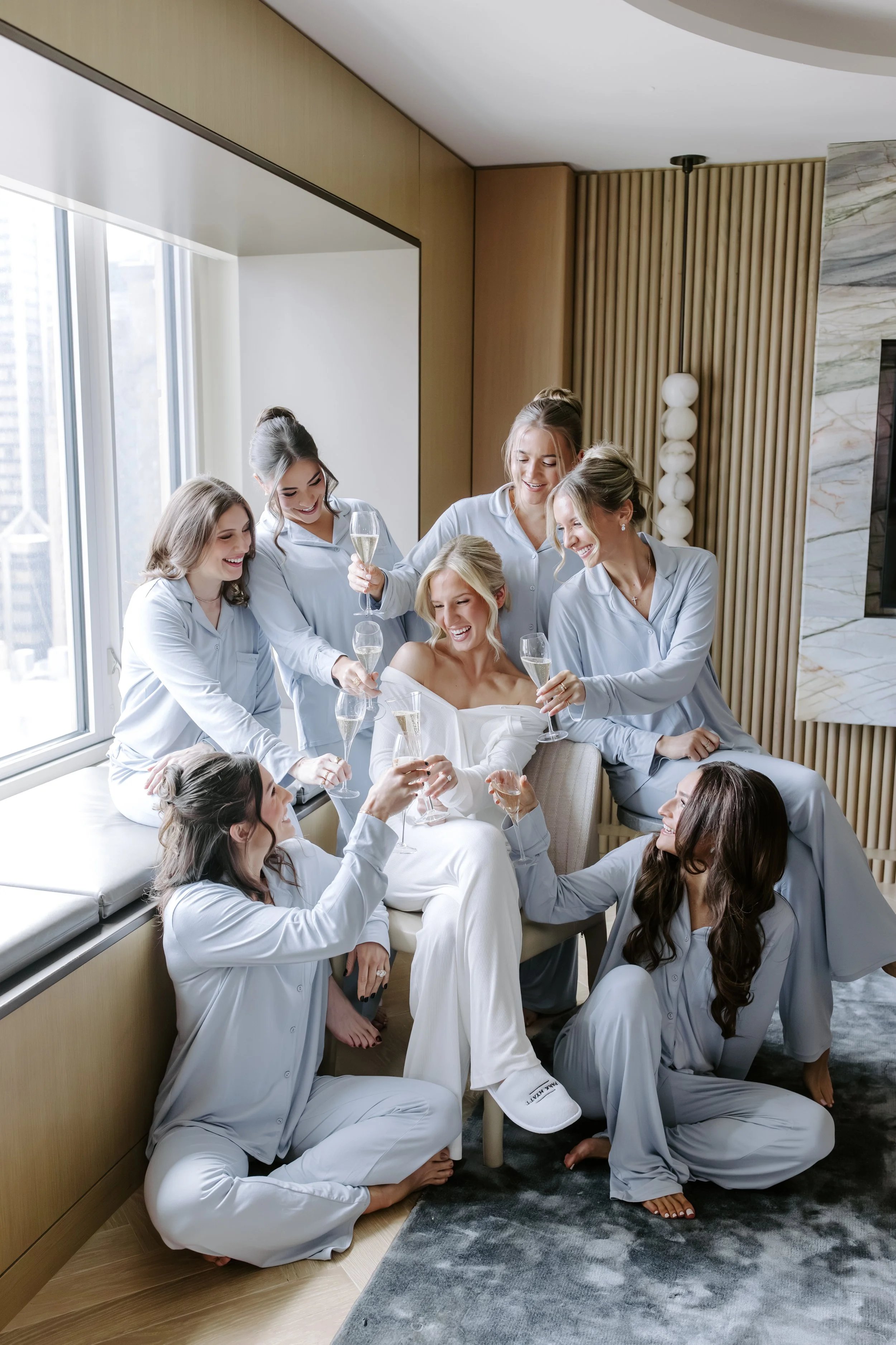 A group of women dressed in matching light blue pajamas celebrating with champagne in a modern living room, some sitting on the floor, others on the couch, smiling, laughing, and toasting.