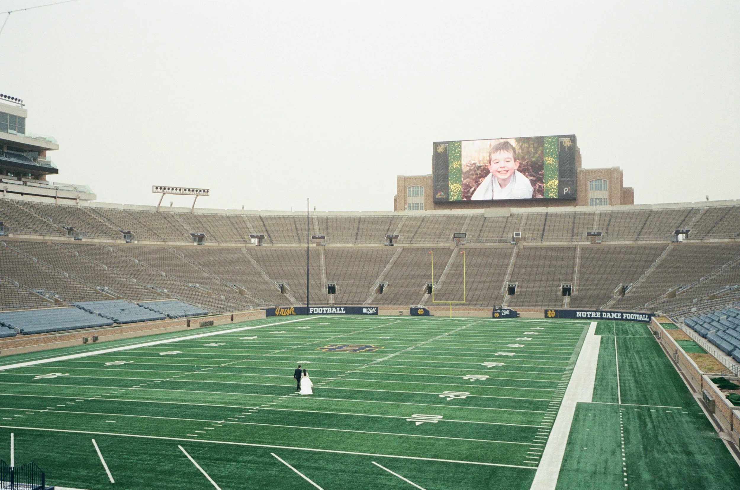 The Notre Dame football stadium empty, with a large digital screen showing a smiling young boy, and a person in a wedding dress walking on the field.