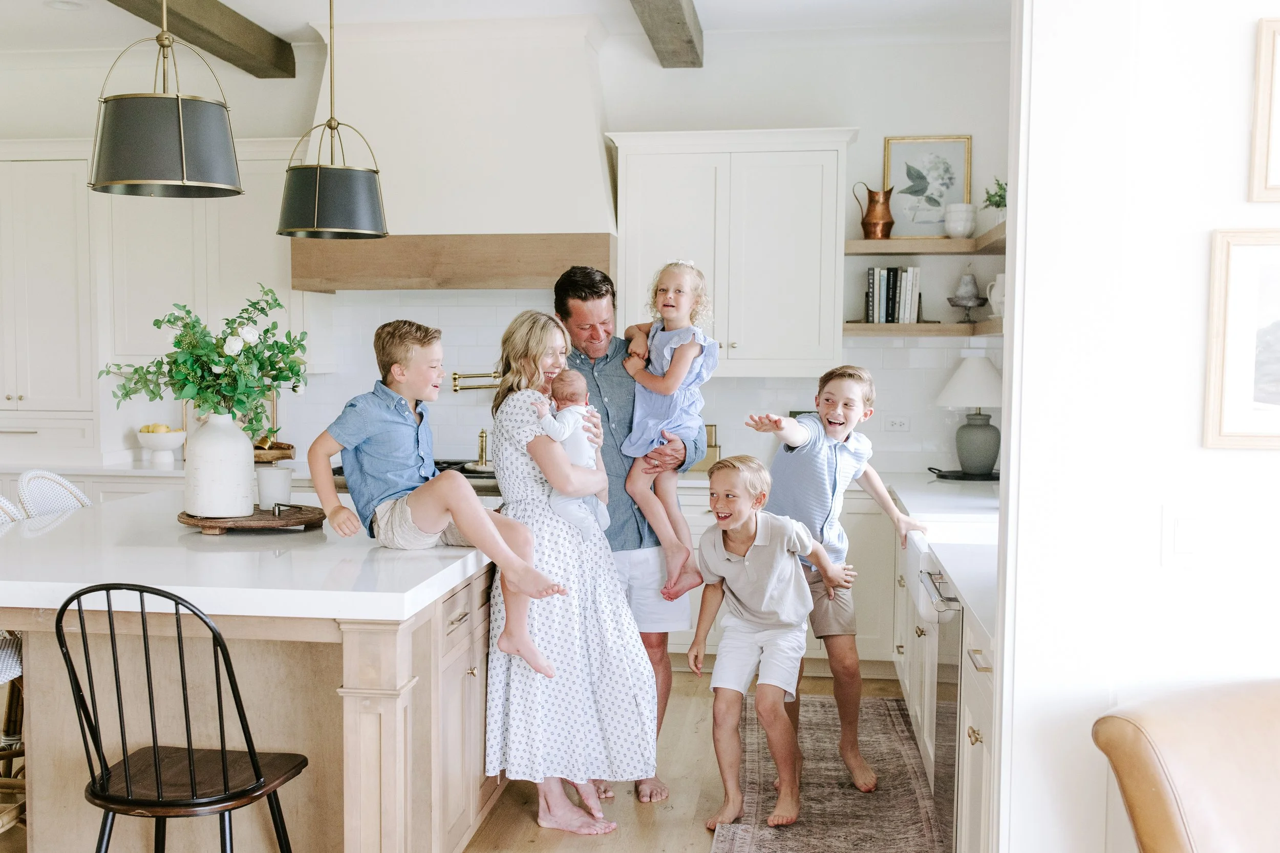 A family enjoying a playful moment in their bright kitchen. A woman is holding a newborn baby while children surround her, laughing and reaching out. The kitchen has white cabinets, a large white countertop with a plant, and natural light filling the