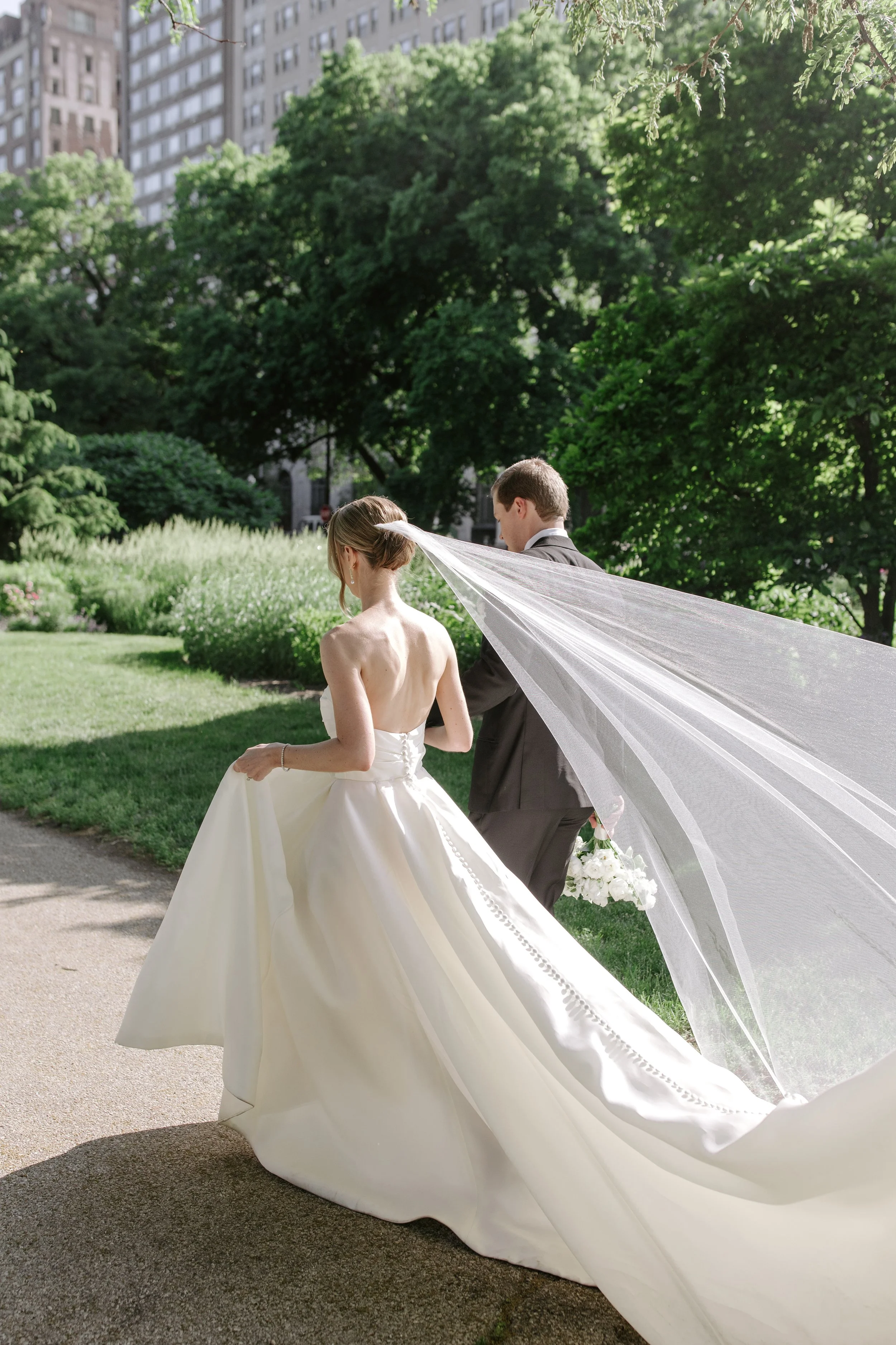 Bride and groom walking together outdoors on a sunny day, surrounded by lush green trees and urban buildings in the background. The bride is in a strapless white wedding gown with a long veil, and the groom is in a dark suit. The bride is holding a b