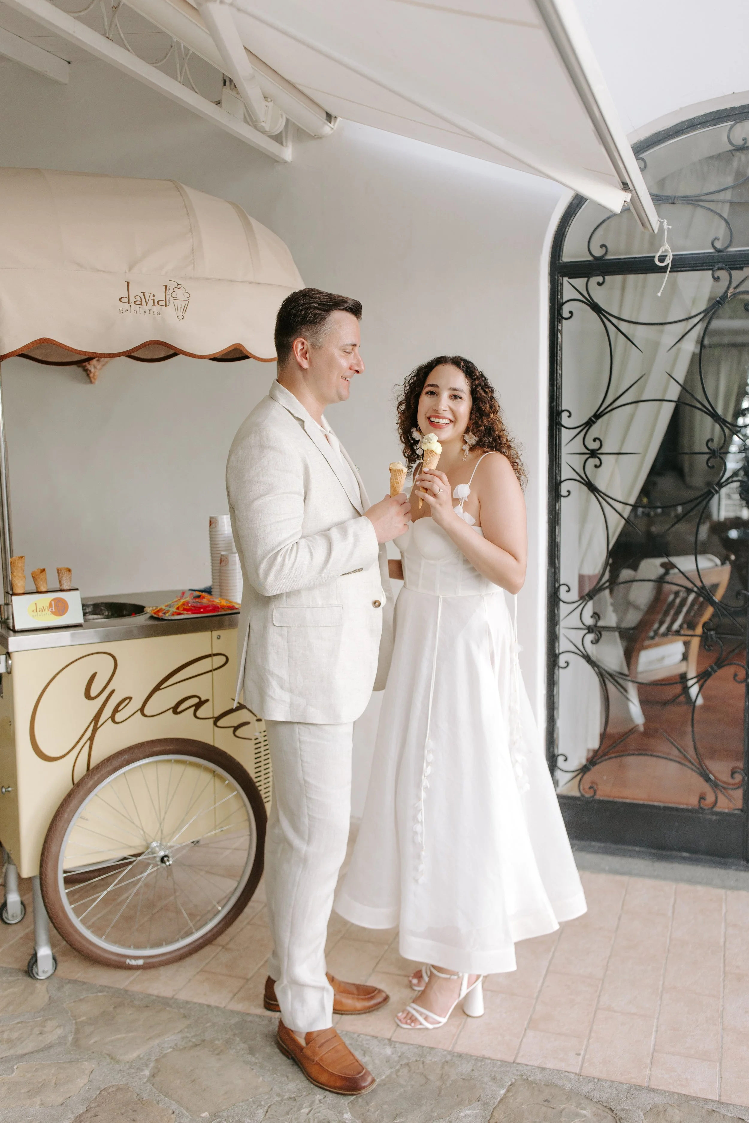 A man and woman in white clothing sharing a moment, holding ice cream cones, in front of a gelato cart named 'DaVinci'.