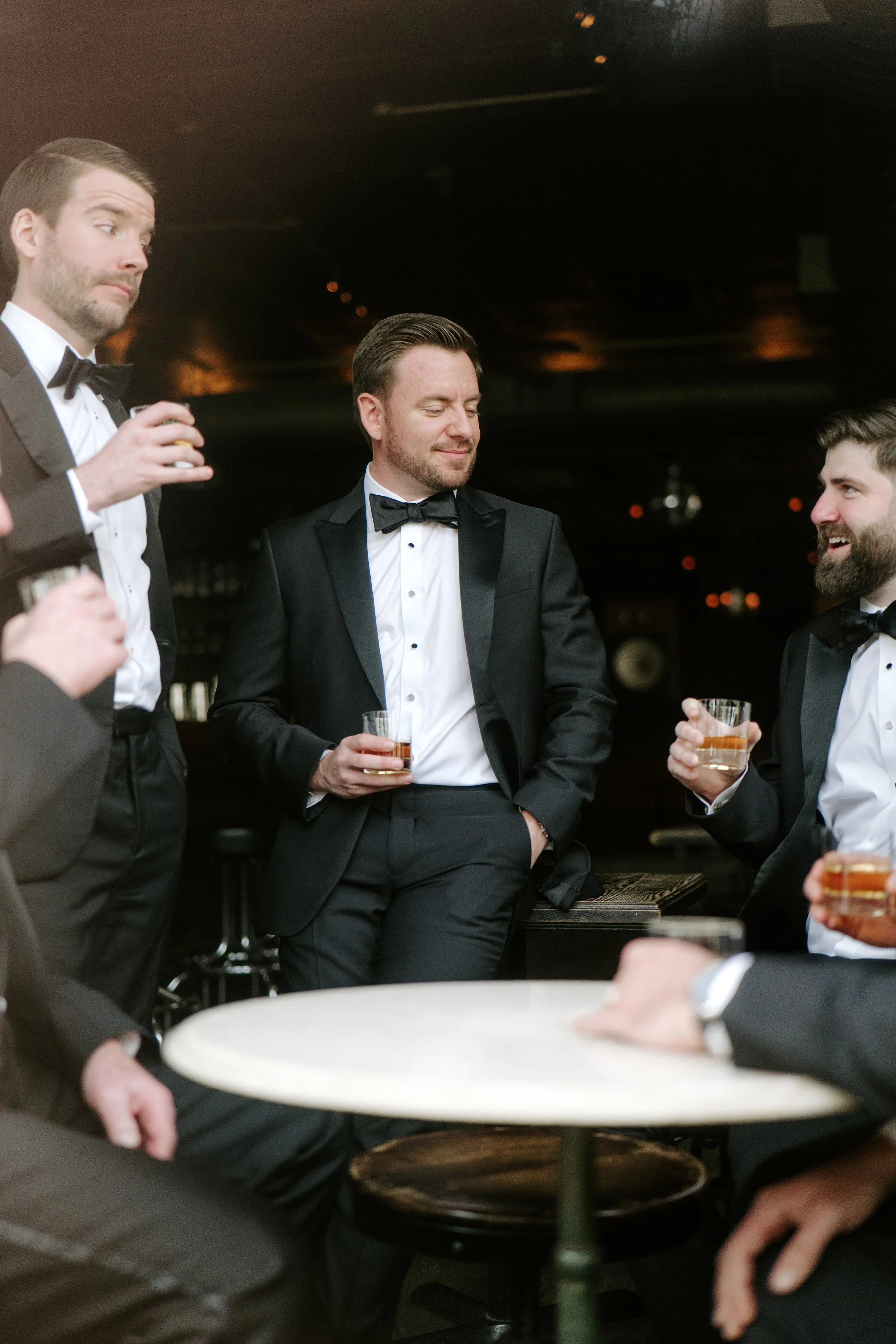 Group of men in tuxedos and bow ties socializing at an elegant event, holding glasses of drinks, in a dimly lit room with a dark ceiling and warm lighting.