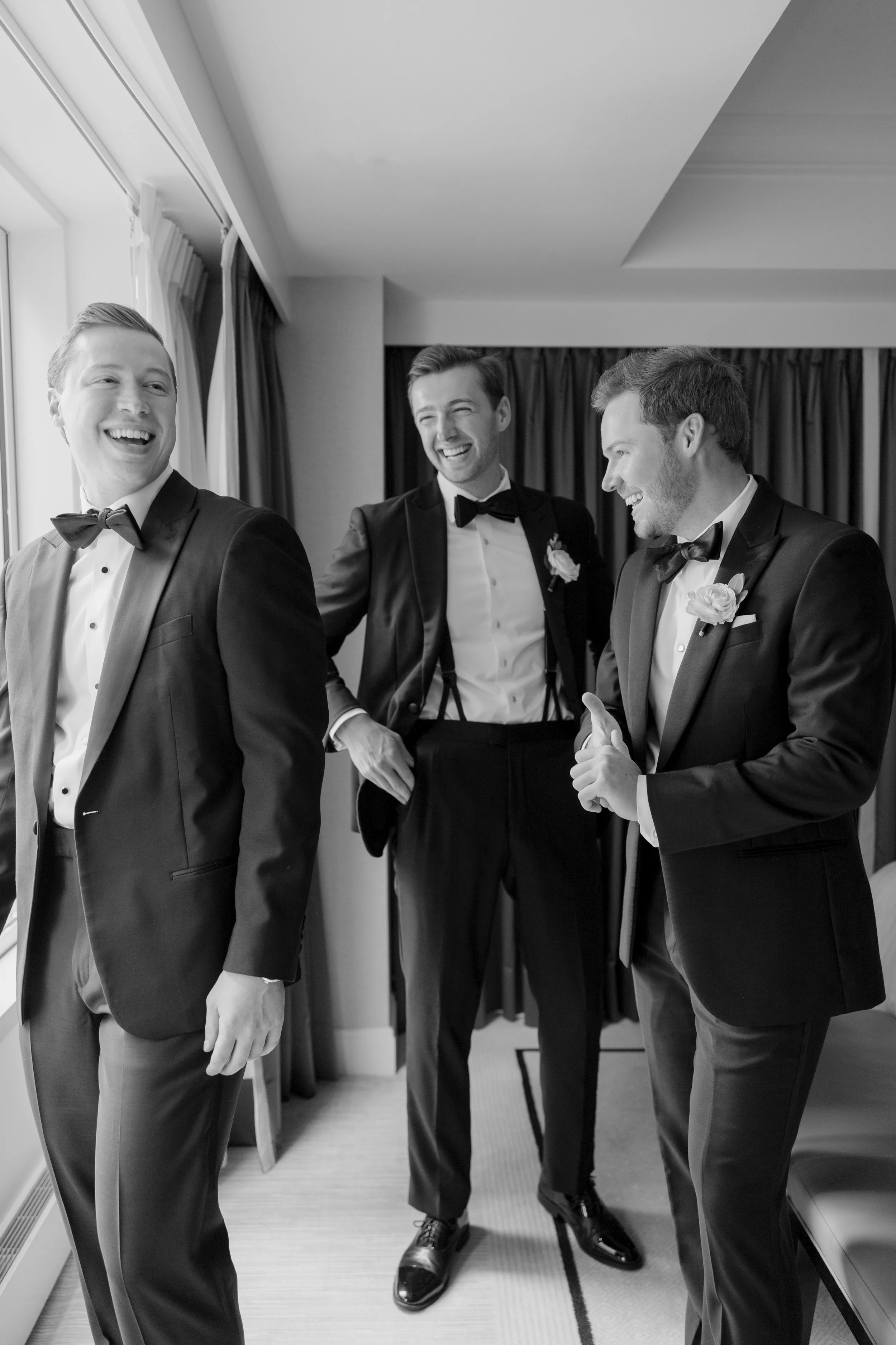 Three men dressed in tuxedos with bow ties, smiling and laughing together in a room with curtains and large windows.