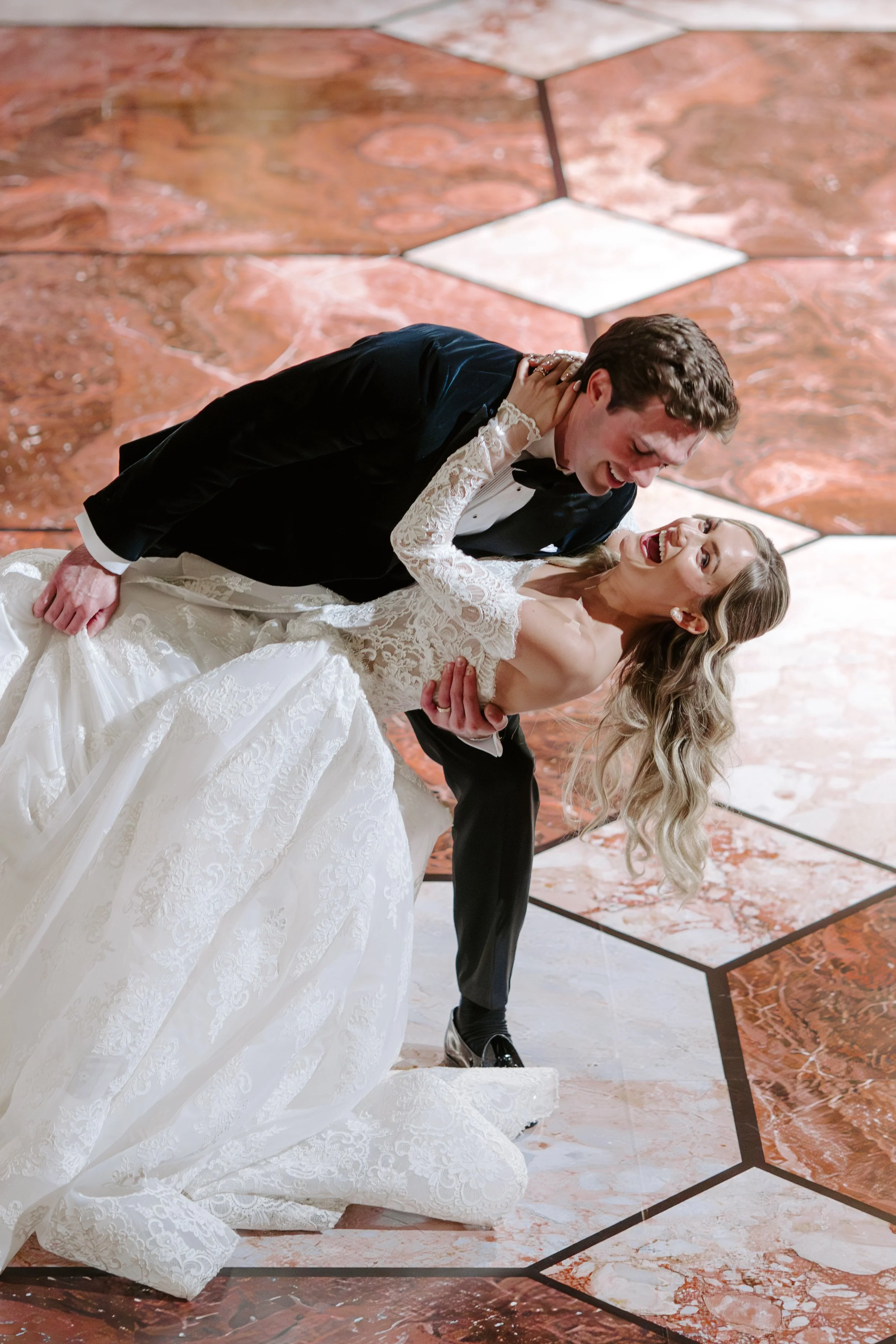 A bride and groom are dancing, with the groom dipping the bride, both smiling and enjoying the moment, on a marble floor.