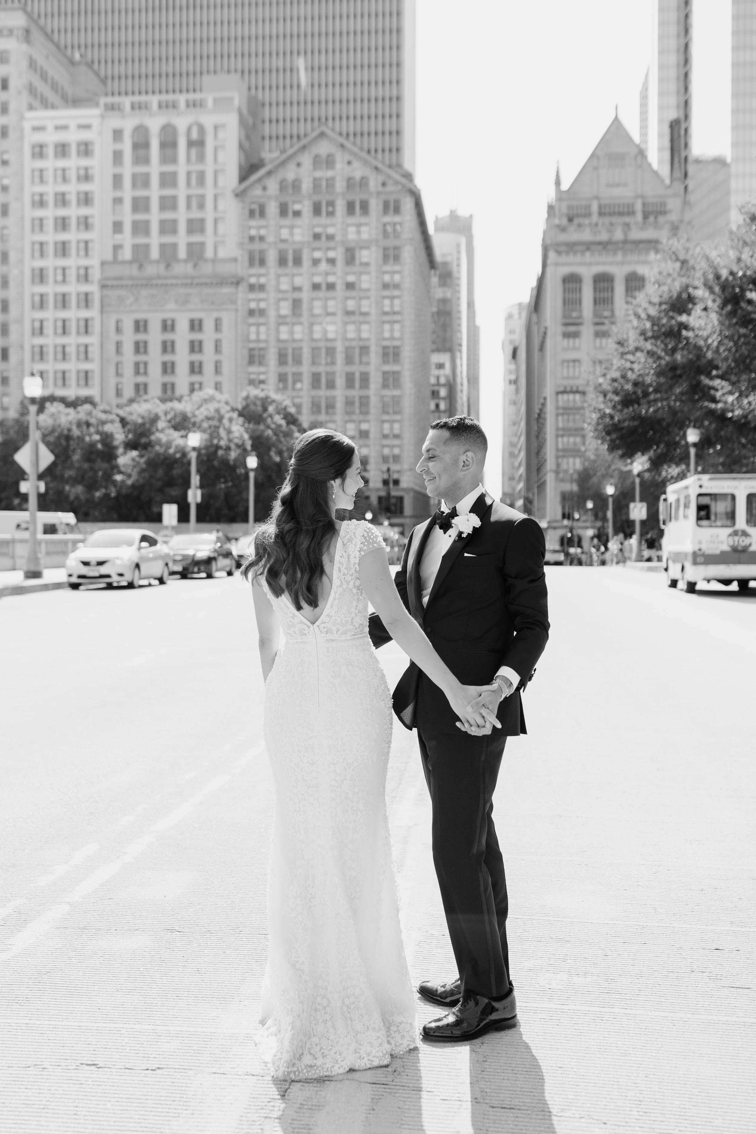A bride and groom holding hands and smiling at each other on a city street with skyscrapers and trees in the background.