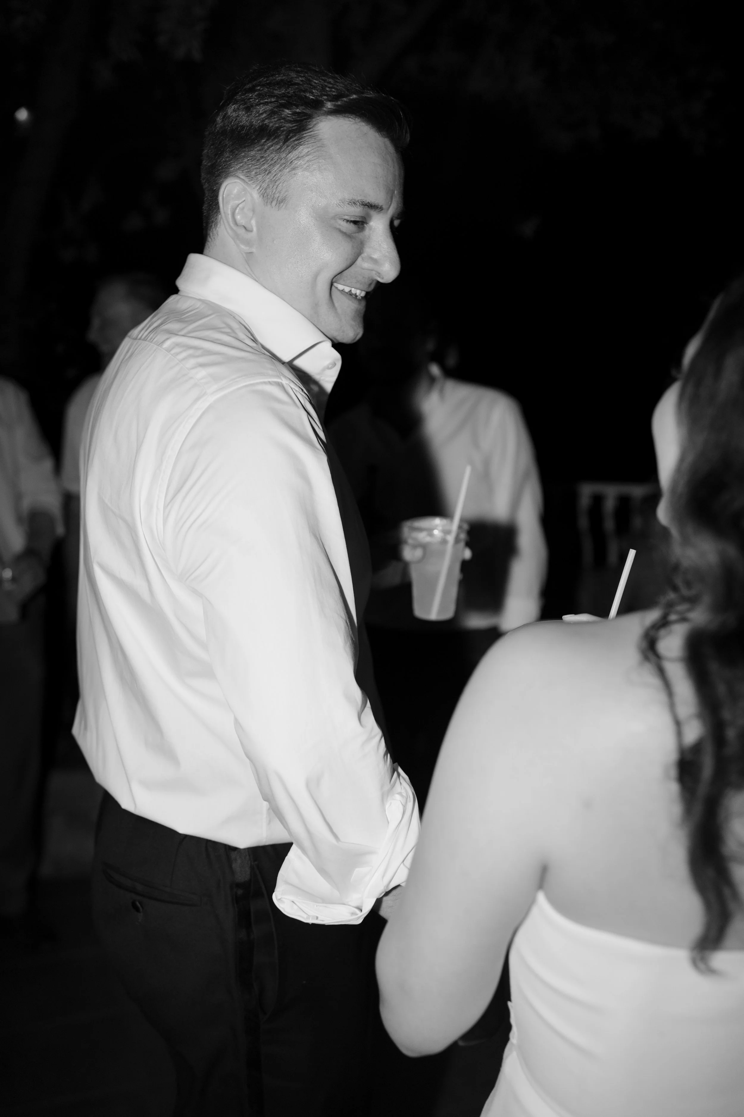 A man in a white shirt smiling while talking to a woman in a sleeveless dress at a social gathering.
