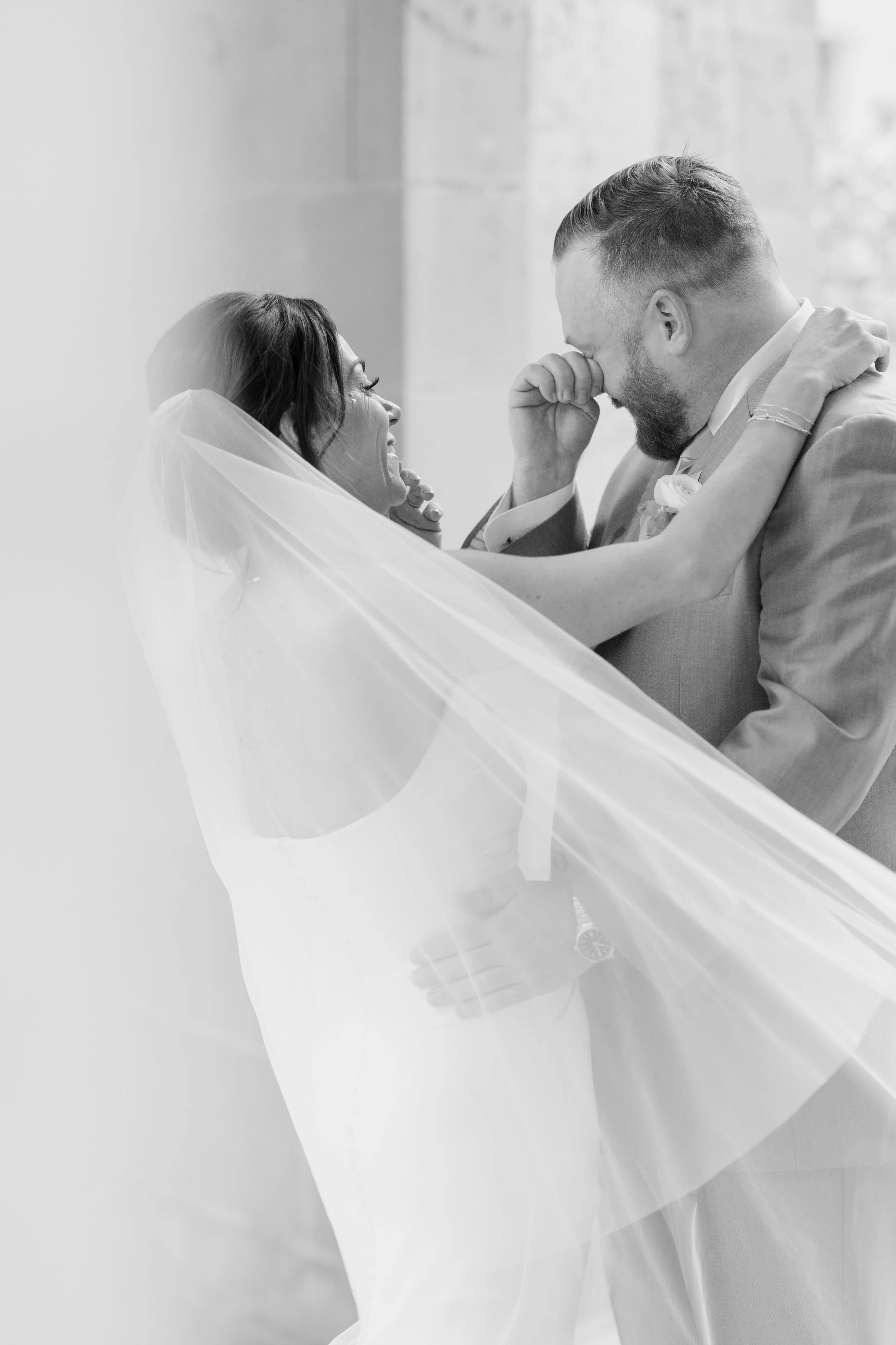 A black and white photo of a bride and groom on their wedding day, smiling and sharing an emotional moment, with the bride wearing a veil and the groom adjusting his suit.