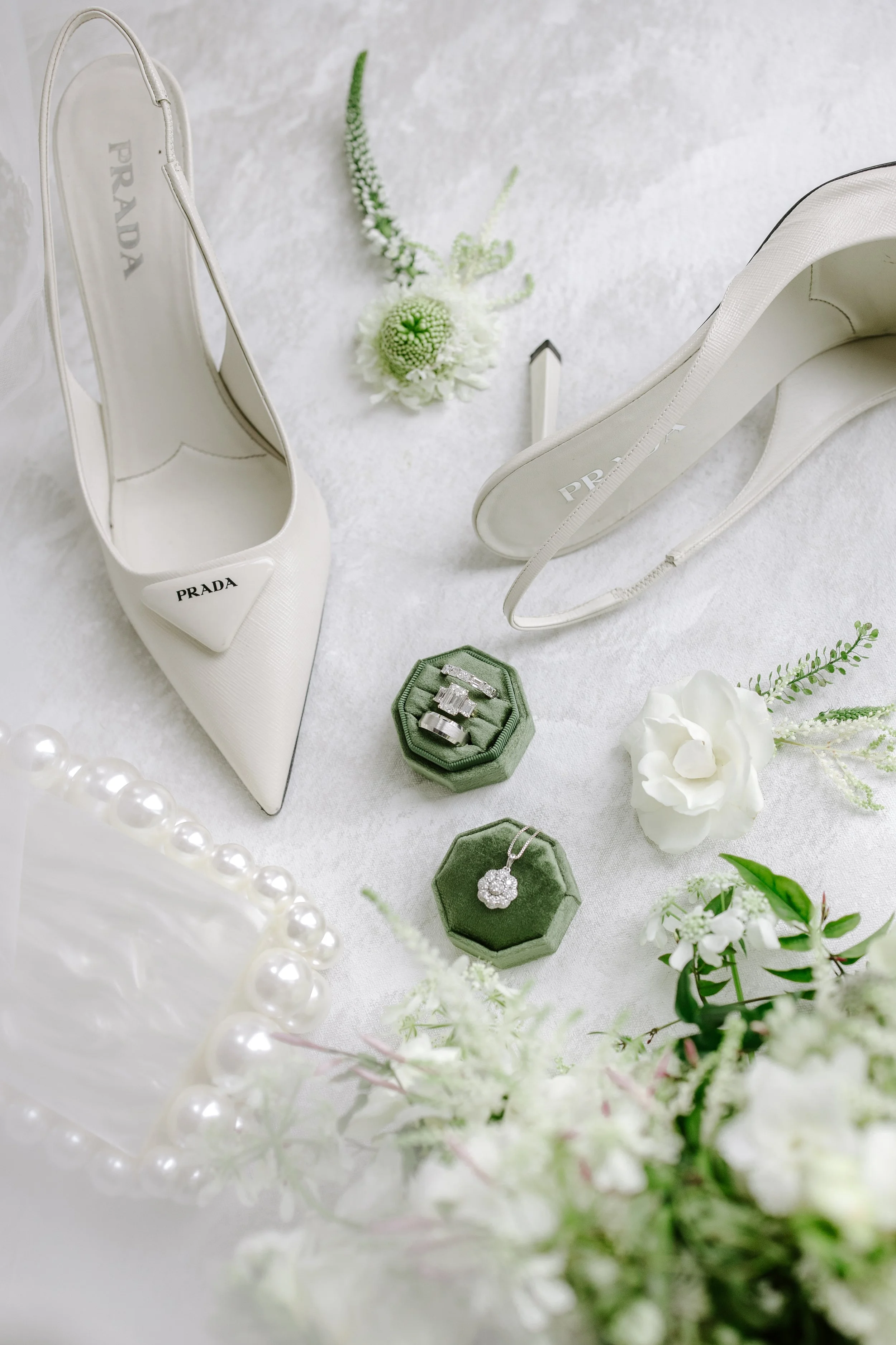 White Prada high-heeled shoes with a bow detail, two green velvet ring boxes with diamond jewelry, white flowers, and a pearl necklace arranged on a white surface.