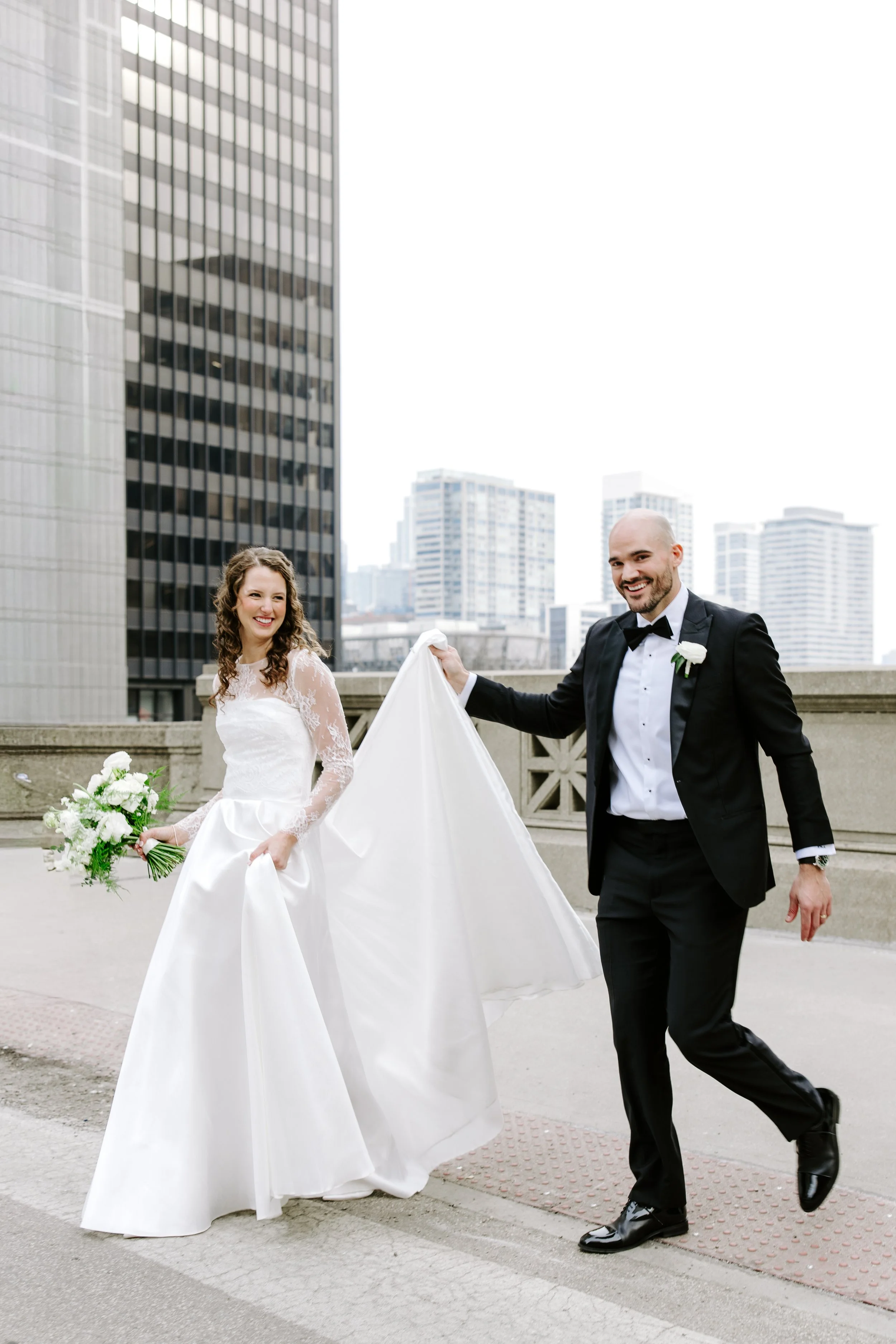 A bride and groom celebrating outdoors on a city rooftop, with tall modern buildings in the background. The bride is holding a bouquet of white flowers and smiling, while the groom, dressed in a black tuxedo, is joyfully holding her train.