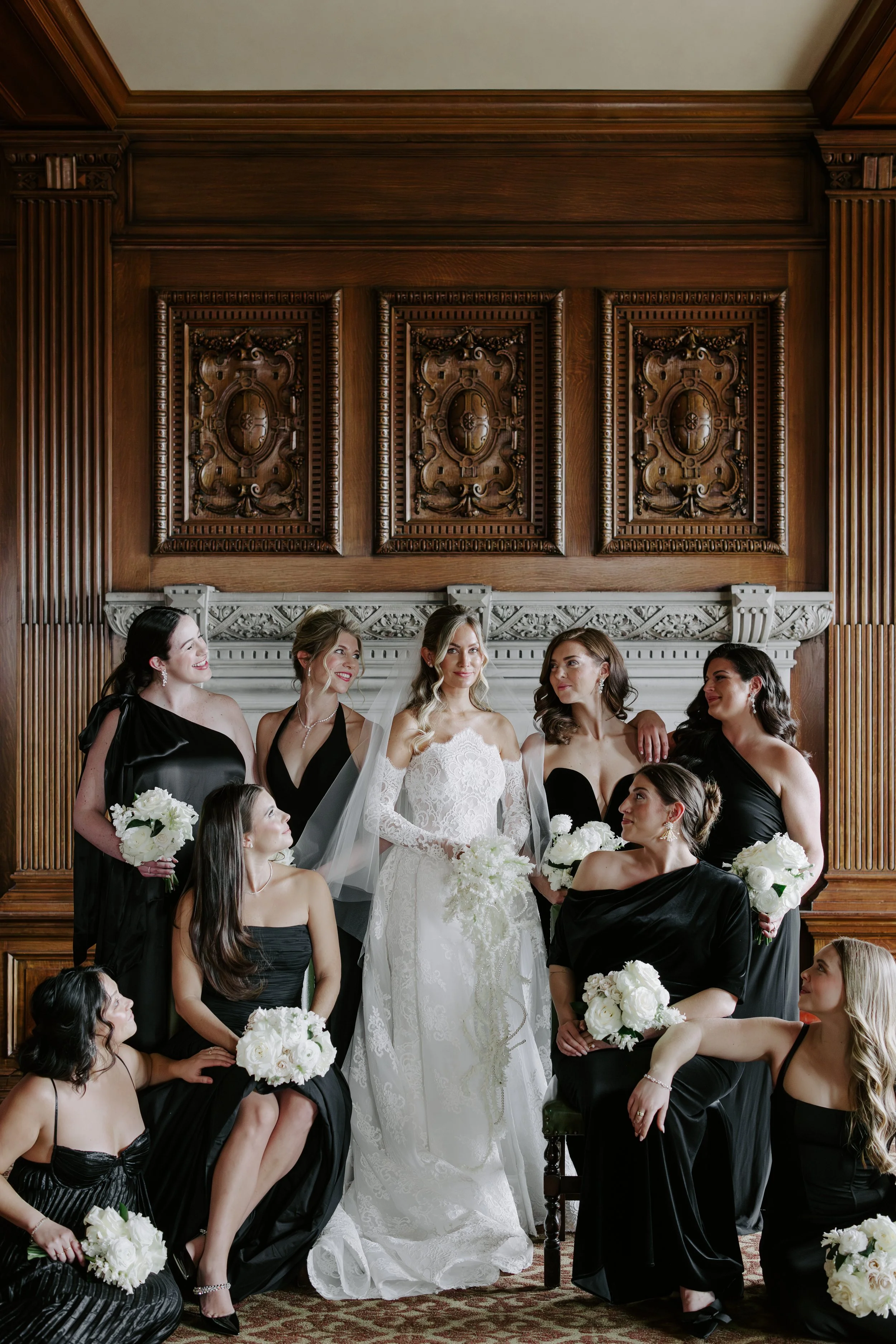 A bride in a white lace wedding dress with long sleeves standing in the center of a group of women dressed in black, holding white bouquets, in an elegant room with dark wood paneling and ornate decor.