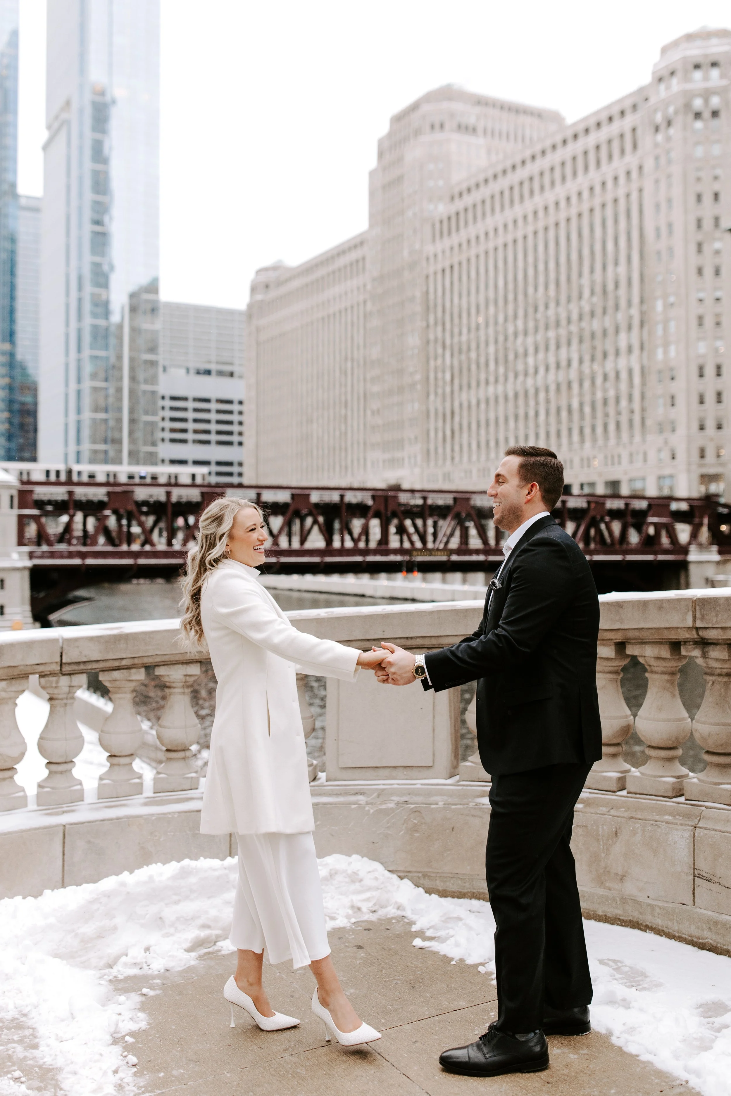 A man and woman standing on a snowy sidewalk in a city, holding hands and smiling at each other. The woman is dressed in a white coat and pants with white high heels, and the man in a black suit and shoes. Tall buildings are visible in the background
