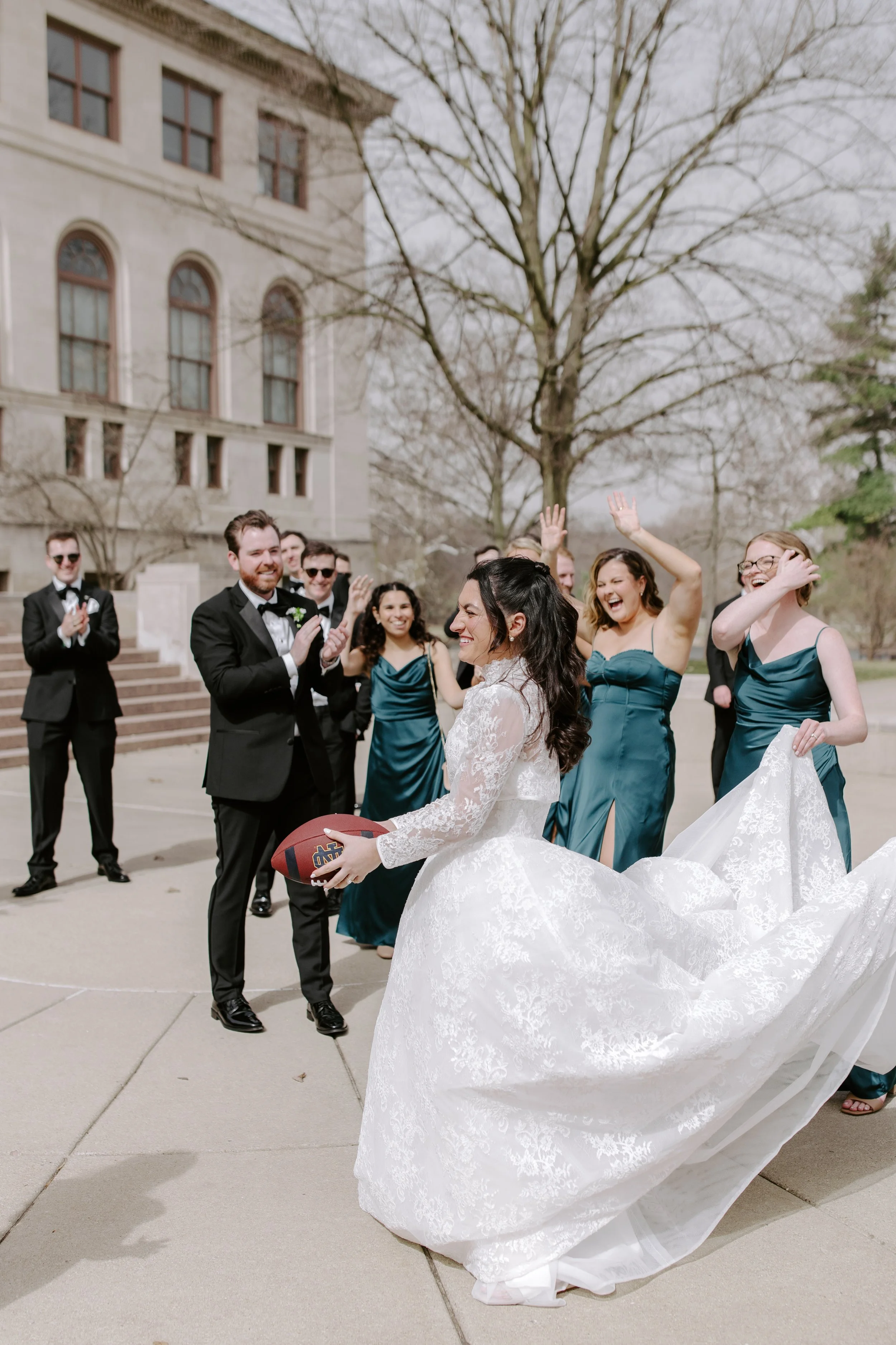 A bride holding a football surrounded by smiling bridesmaids and groomsmen outside a historic building with leafless trees in the background.