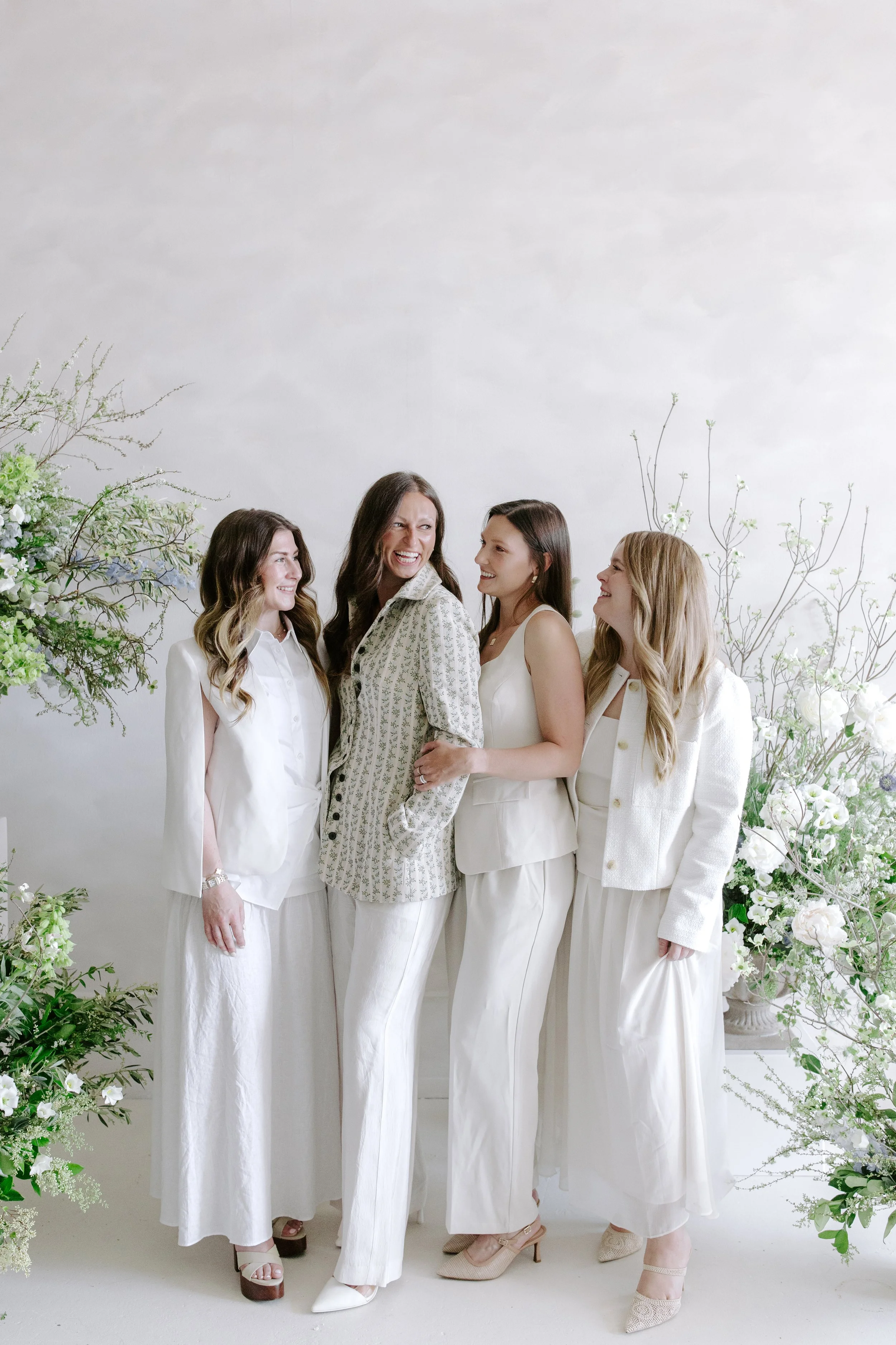 Four women standing together, dressed in white outfits, smiling and looking at each other in front of a white wall decorated with white flowers and greenery.