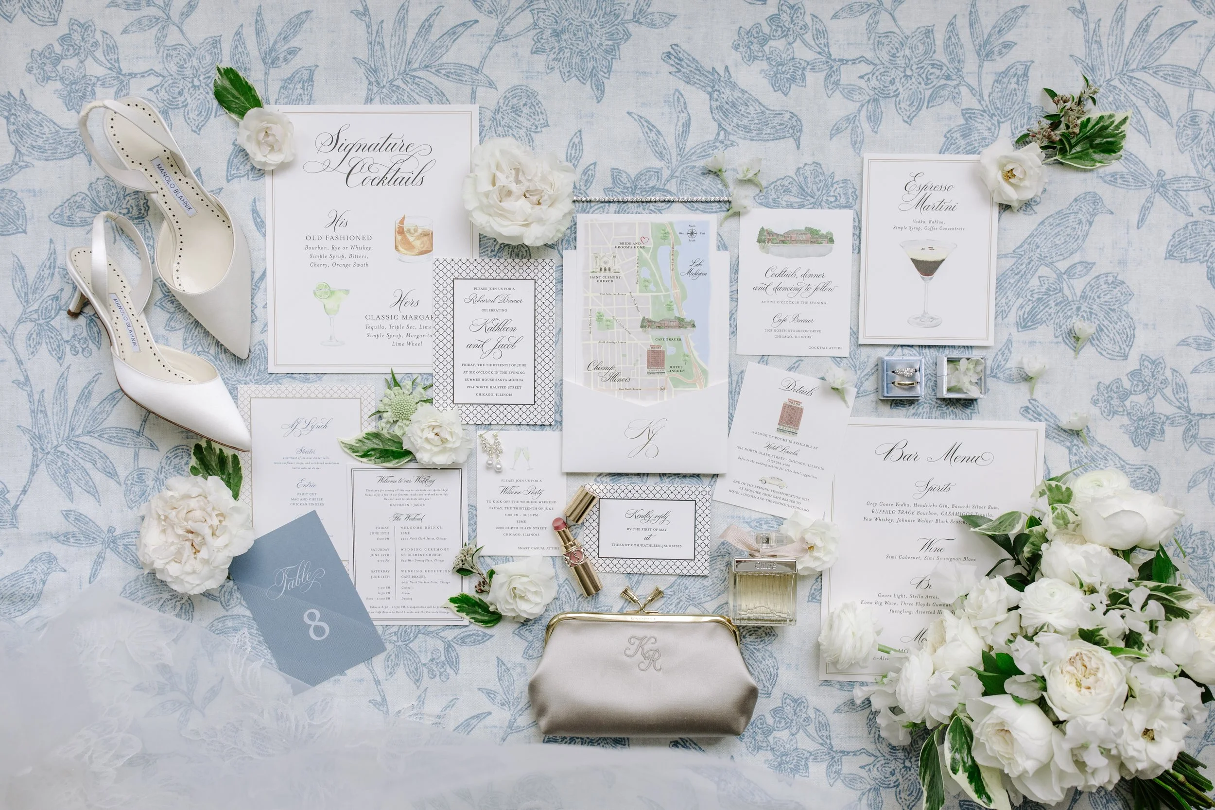 Wedding invitation suite on a floral patterned tablecloth, decorated with white flowers, a pair of white high heels, and various stationery including a map, menu, and place cards.