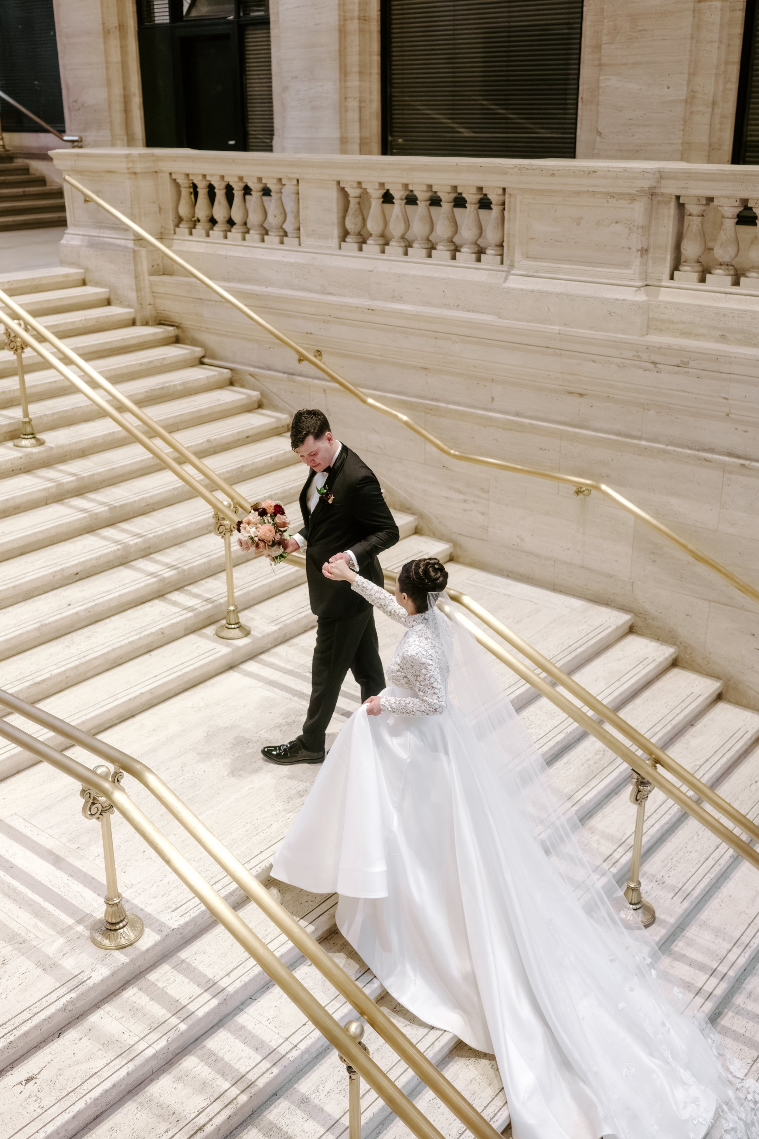 A bride and groom ascending a grand marble staircase inside a building, with the bride wearing a white wedding dress and the groom in a black tuxedo, holding a bouquet of flowers.
