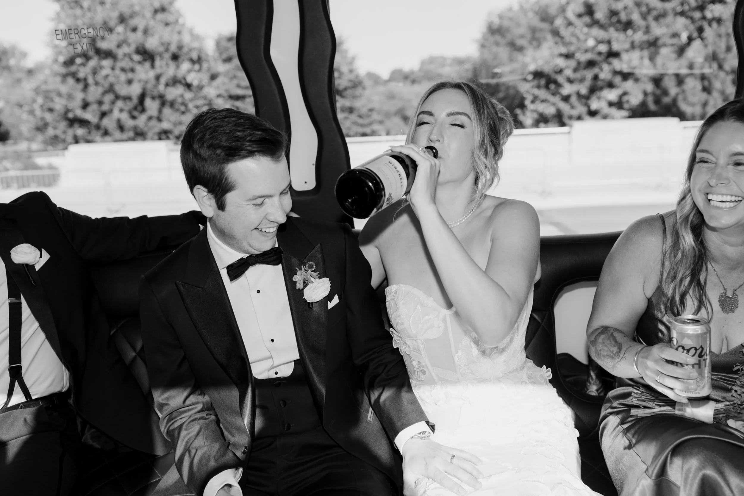 A black and white photo of a wedding celebration with three people sitting in a car. The bride is drinking from a bottle, the groom is smiling, and a woman to the right is holding a drink and smiling.