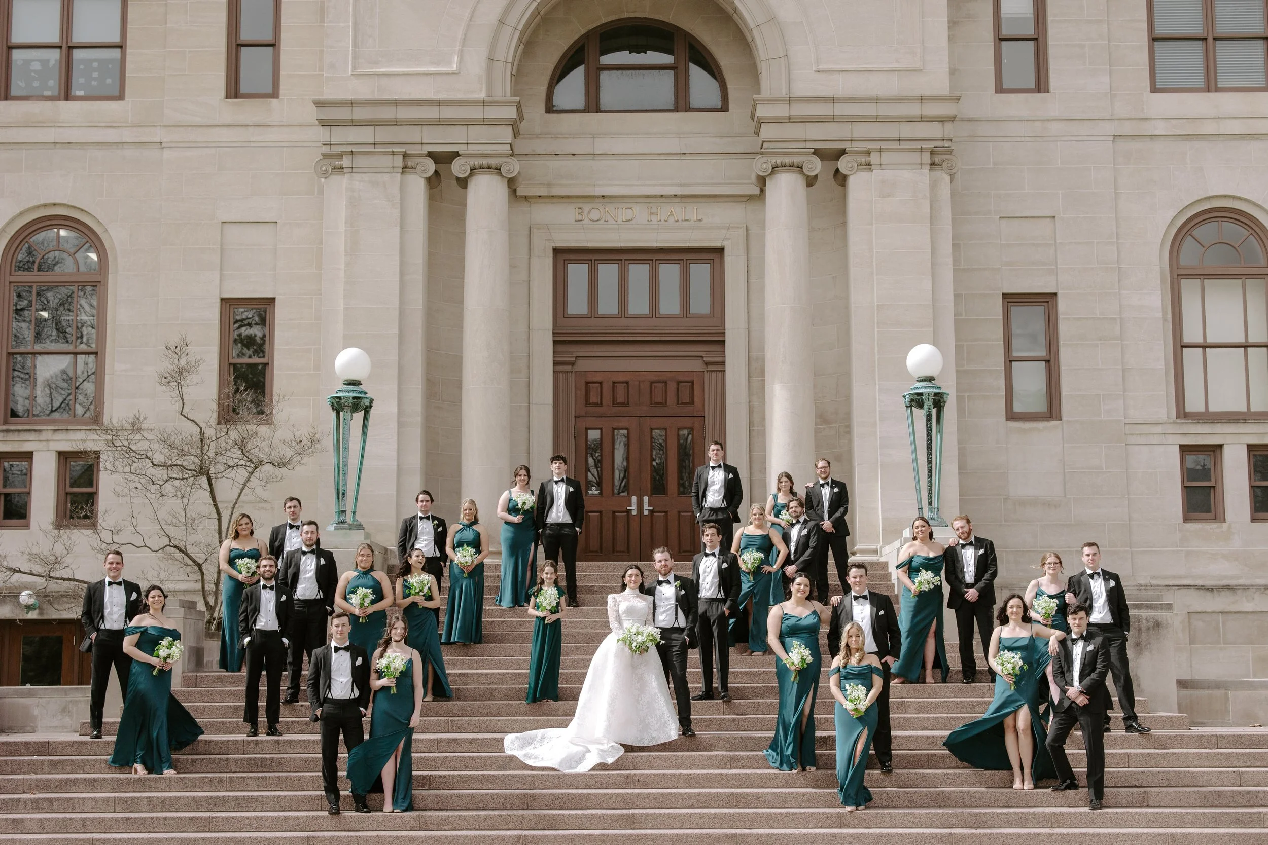 A group of wedding party members standing on the steps of a stone building labeled 'Bond Hall,' with bridesmaids in teal dresses and groomsmen in tuxedos, posing for a photograph.