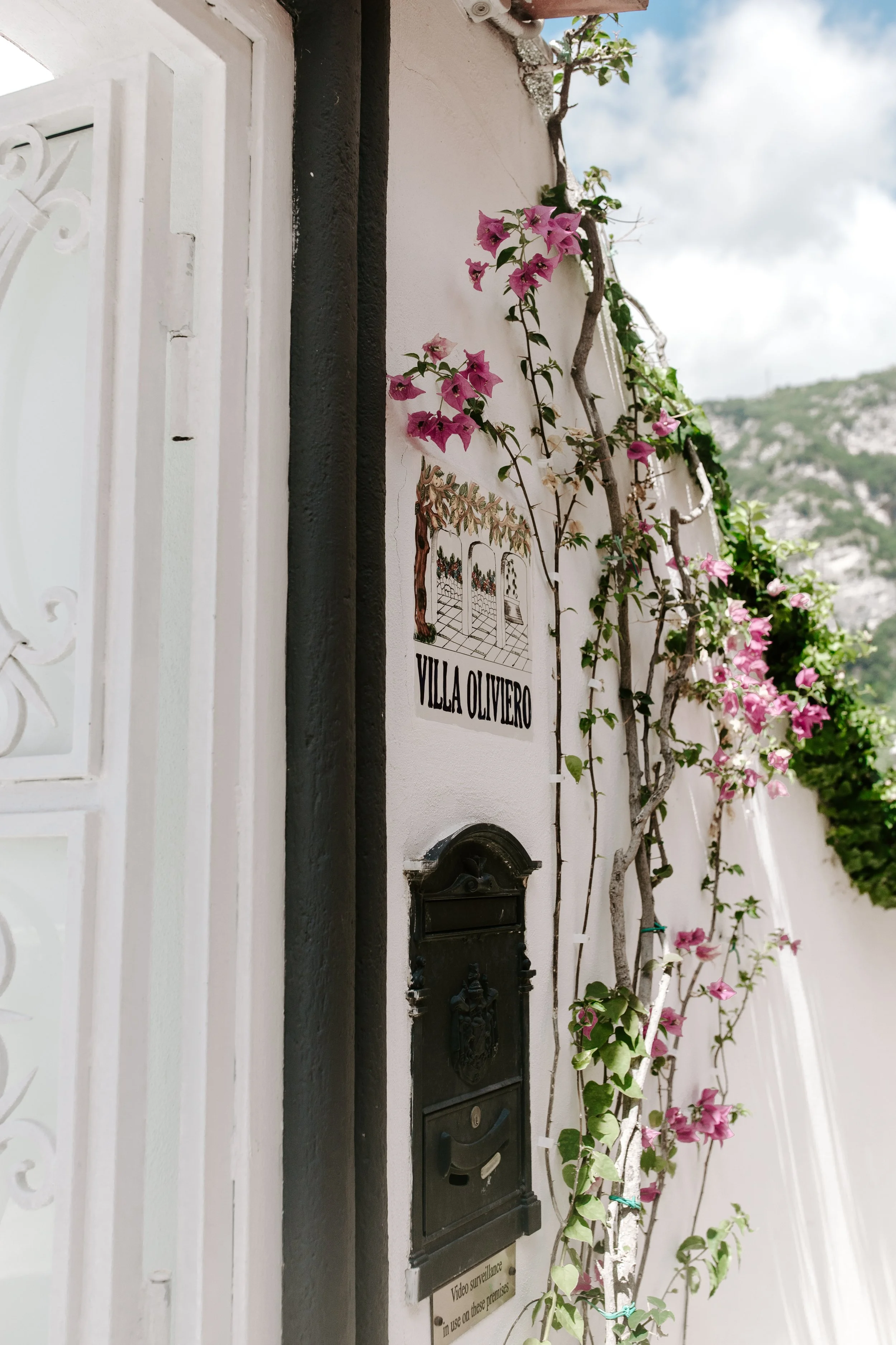 A white wall with a sign that reads "Villa Olivero" and a small illustrated garden scene. There is a black mailbox and a mailbox slot below the sign. Pink bougainvillaea flowers and green vines grow along the wall, with a mountain landscape in the ba