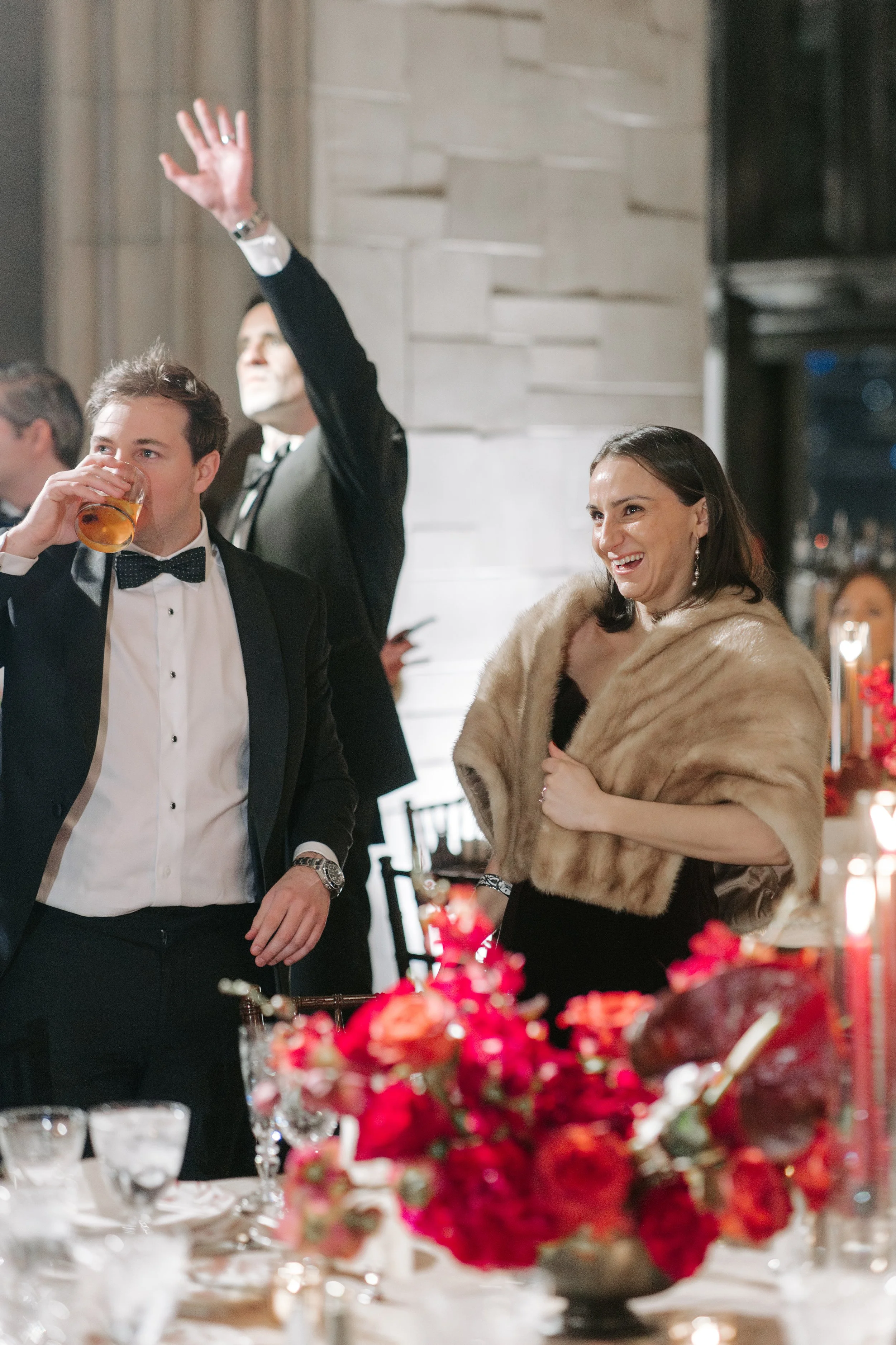 People at a formal event, a man in a tuxedo drinking, a woman in a fur stole smiling, and a man in a suit with his hand raised.