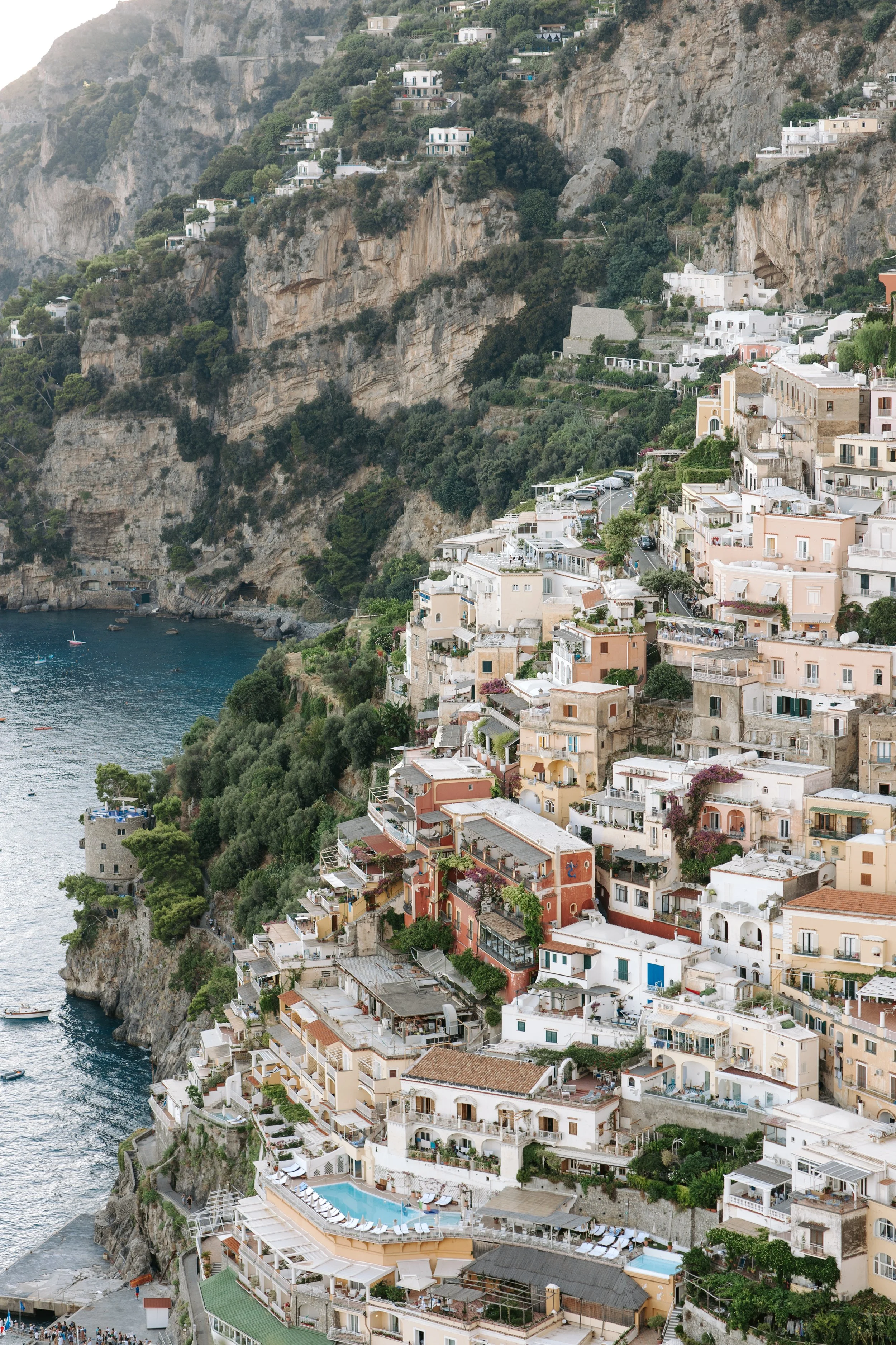 A hillside village on a coast with white and pastel-colored buildings, some with terraces and balconies, winding roads, and lush greenery, overlooking the sea with small boats. There is a mix of residential dwellings, some with swimming pools, and st