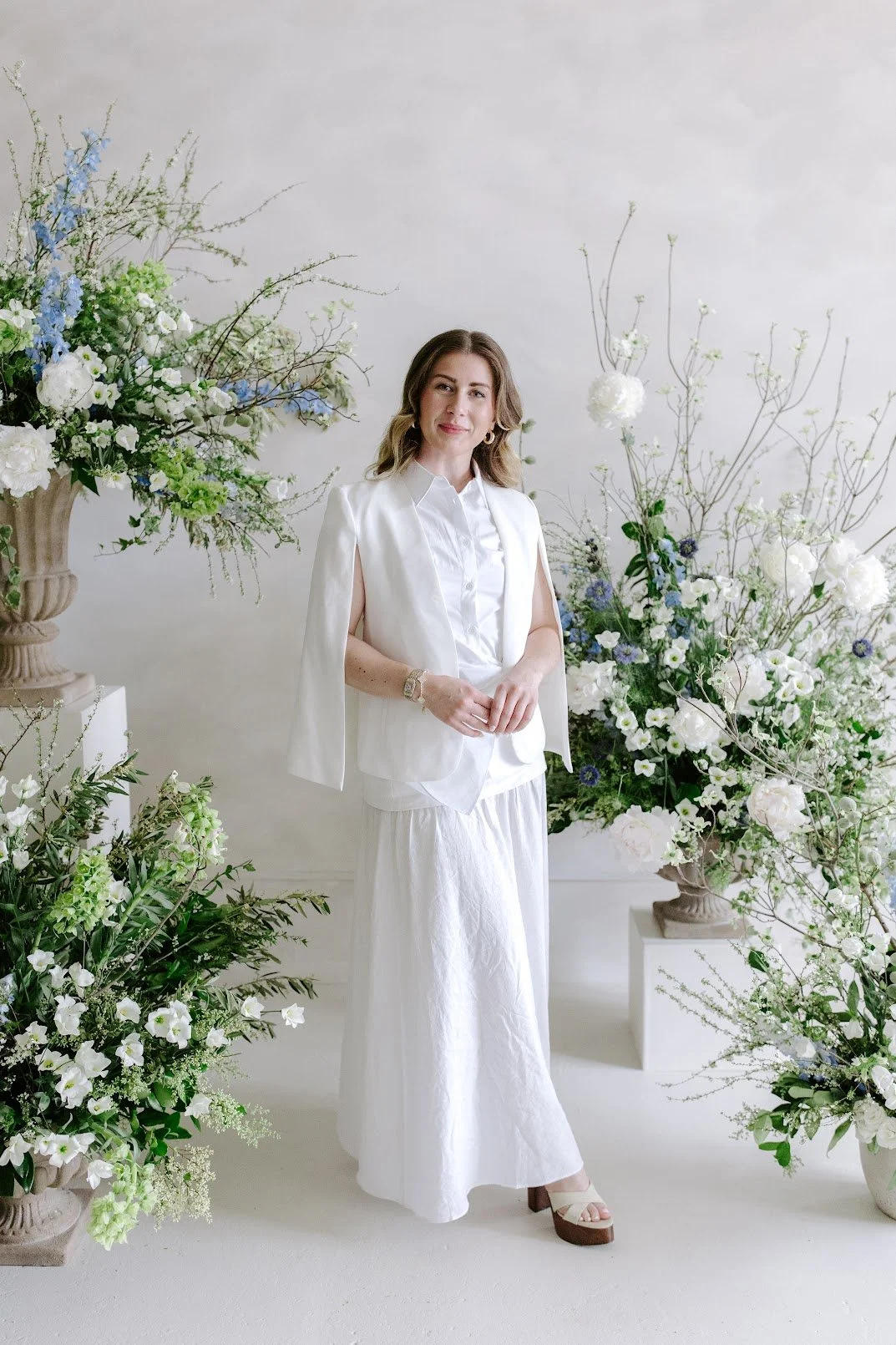 Woman wearing white outfit standing amidst a display of large floral arrangements in a neutral-toned setting.