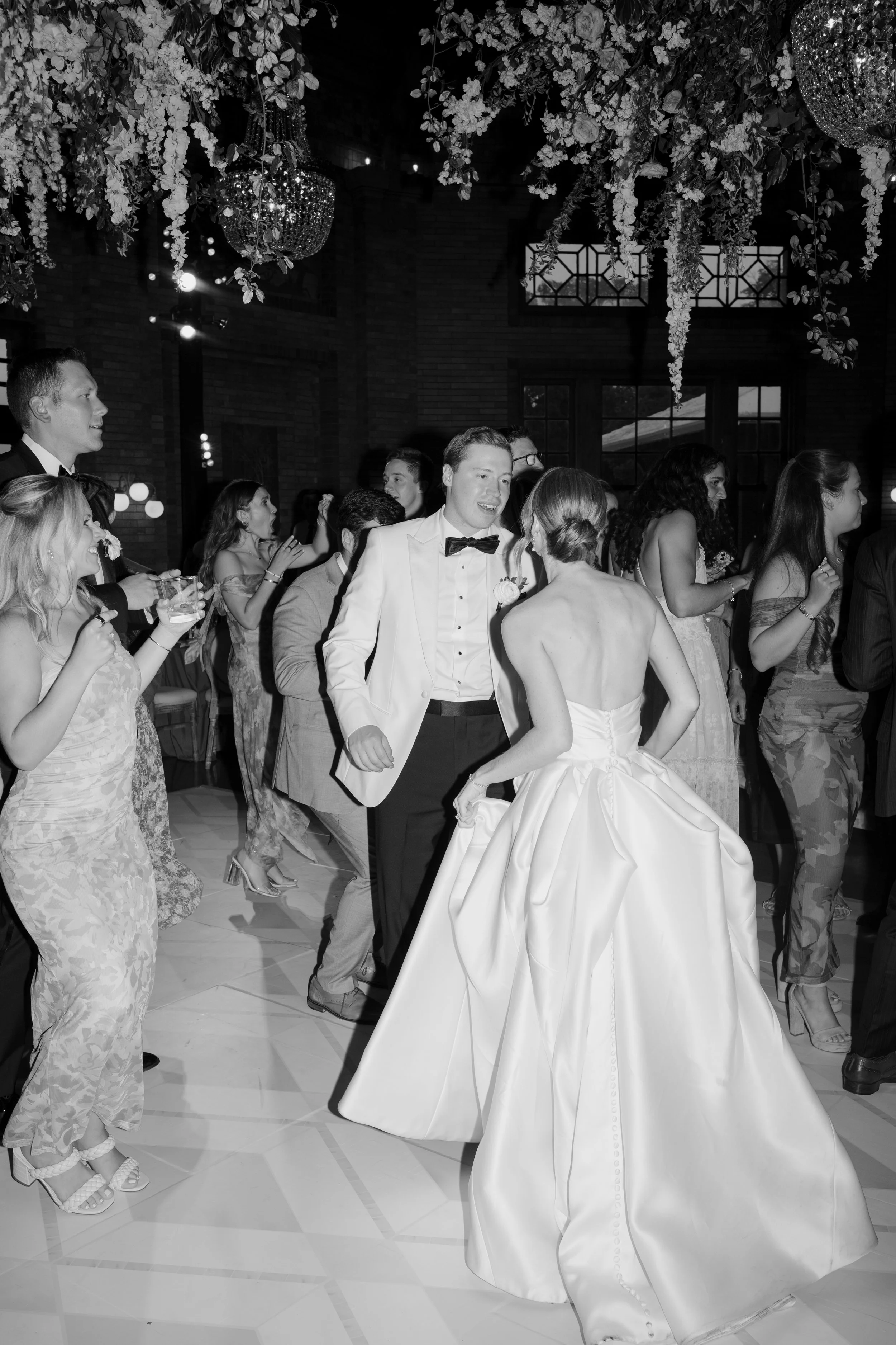 A bride and groom dancing at their wedding reception, surrounded by guests, with floral decorations hanging from the ceiling and large windows in the background.