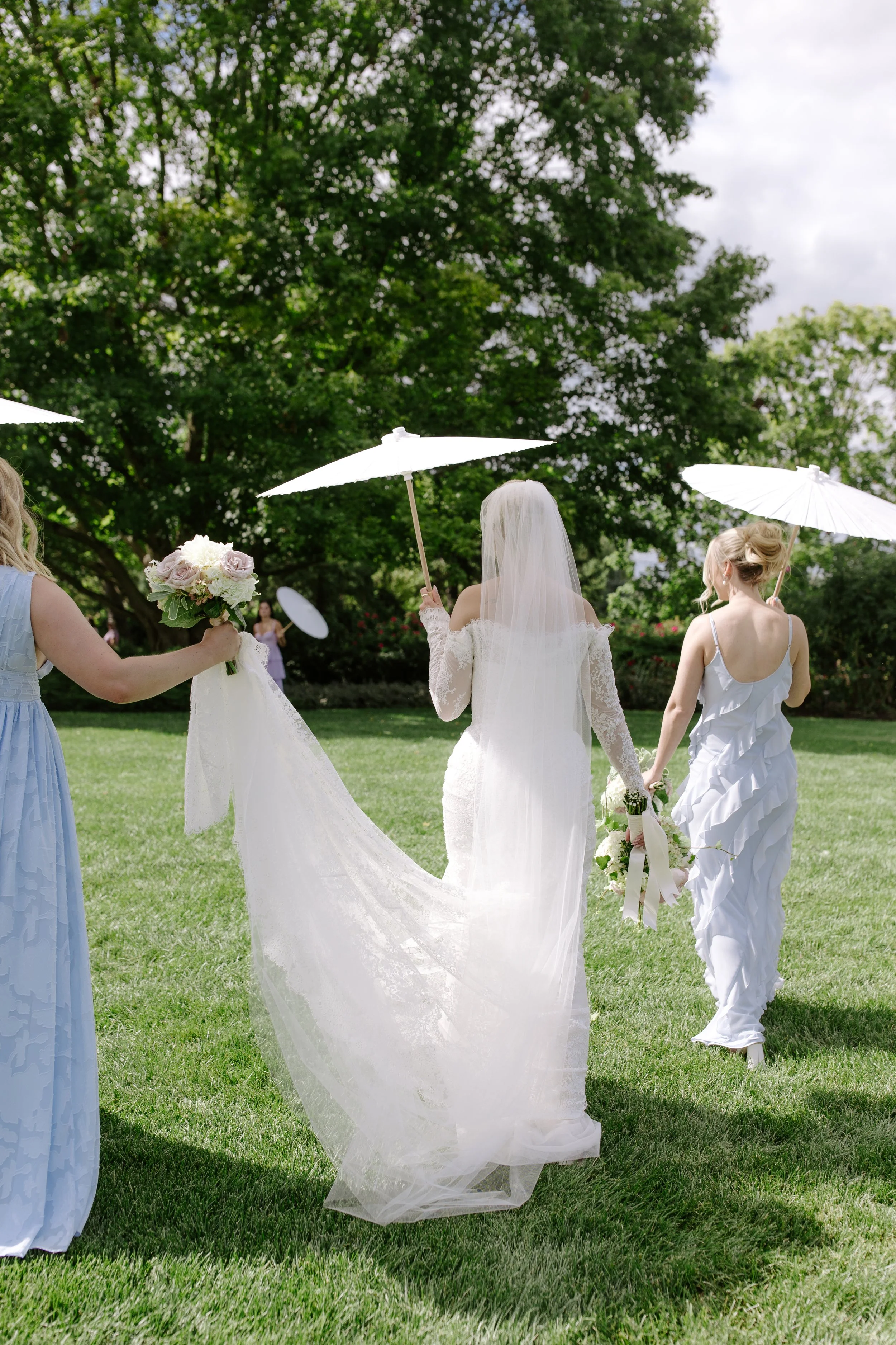 A bride with a veil holding a bouquet of flowers and two women holding white umbrellas walking on a grassy field during a wedding ceremony outdoors, with large trees in the background.