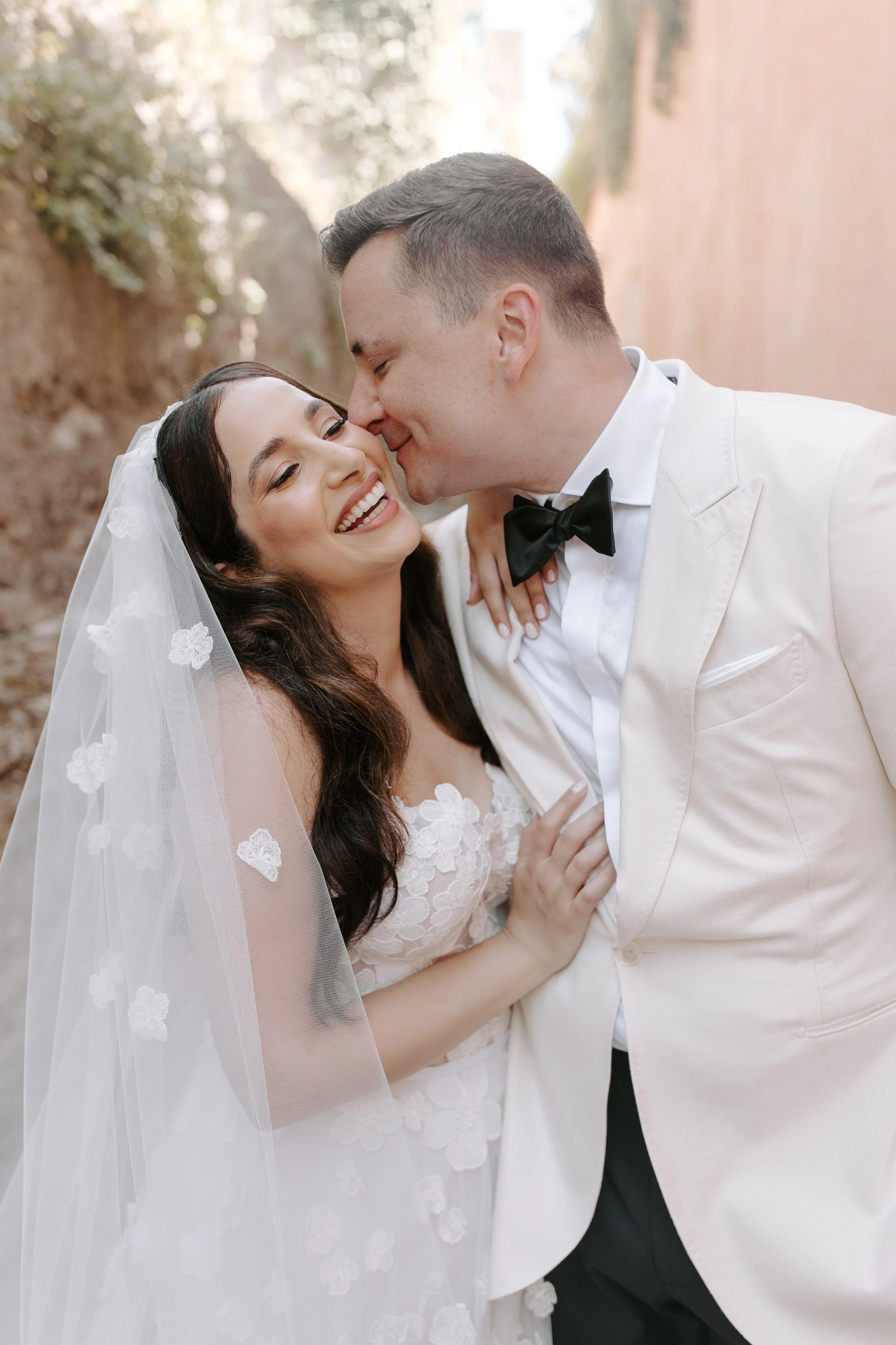 Bride and groom sharing a kiss outdoors on their wedding day, with the bride wearing a lace wedding dress and veil, and the groom in a white tuxedo with black bow tie.