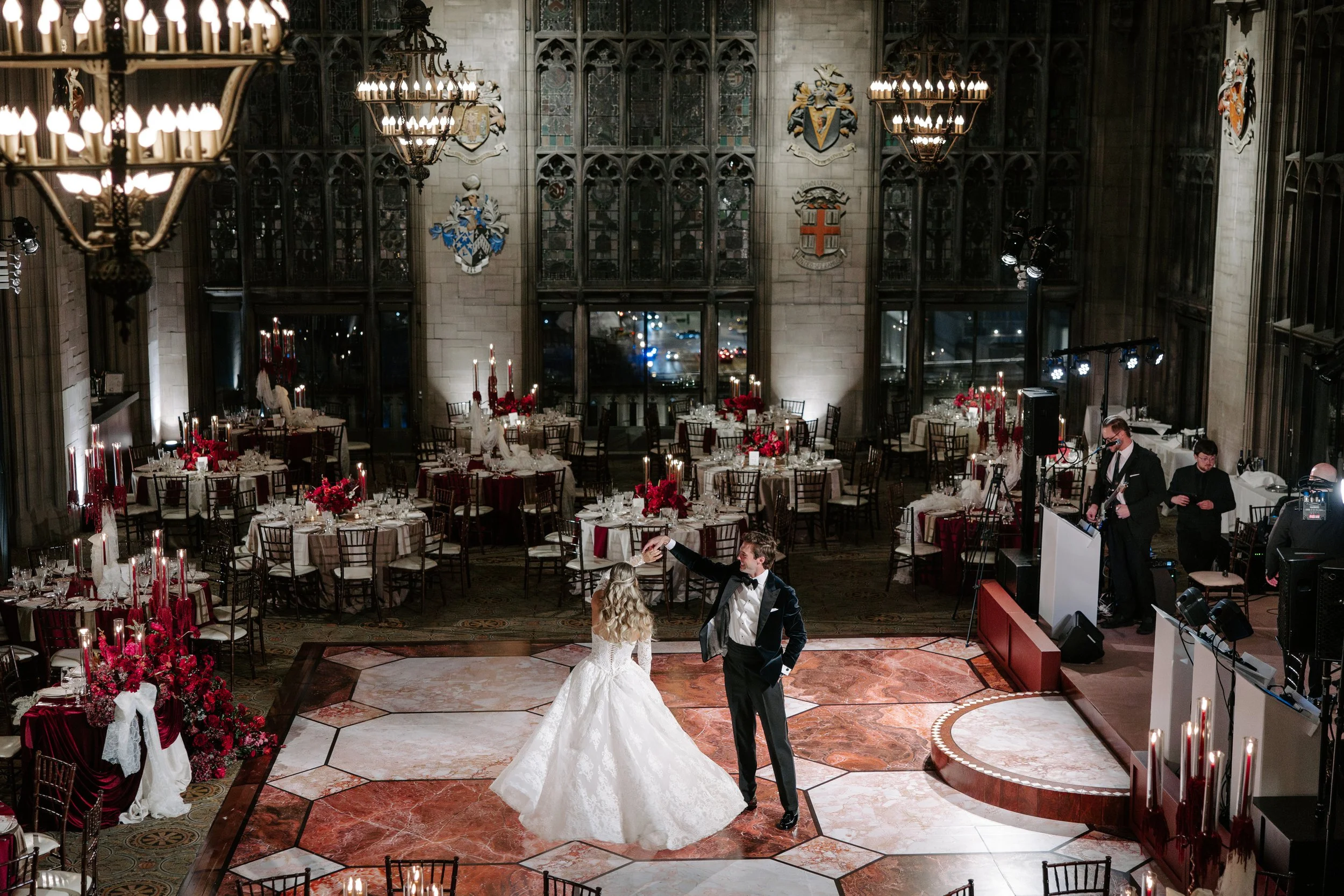 A wedding reception held in a grand hall with high ceilings and large stained glass windows. A bride and groom are dancing on the floor, surrounded by round tables decorated with red floral centerpieces and lit candles. There are chandeliers hanging 