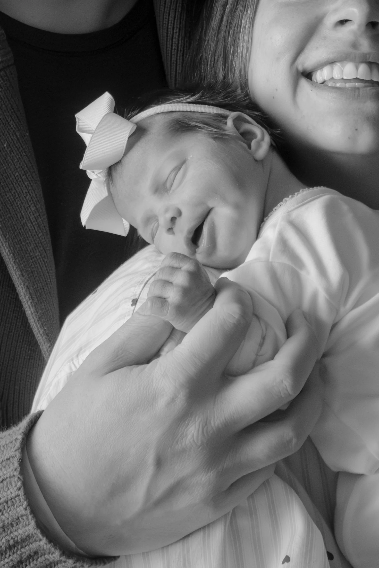 A woman smiling with her eyes closed holding a sleeping baby close to her chest. The baby is wearing a headband with a bow and appears to be very young, possibly a newborn.