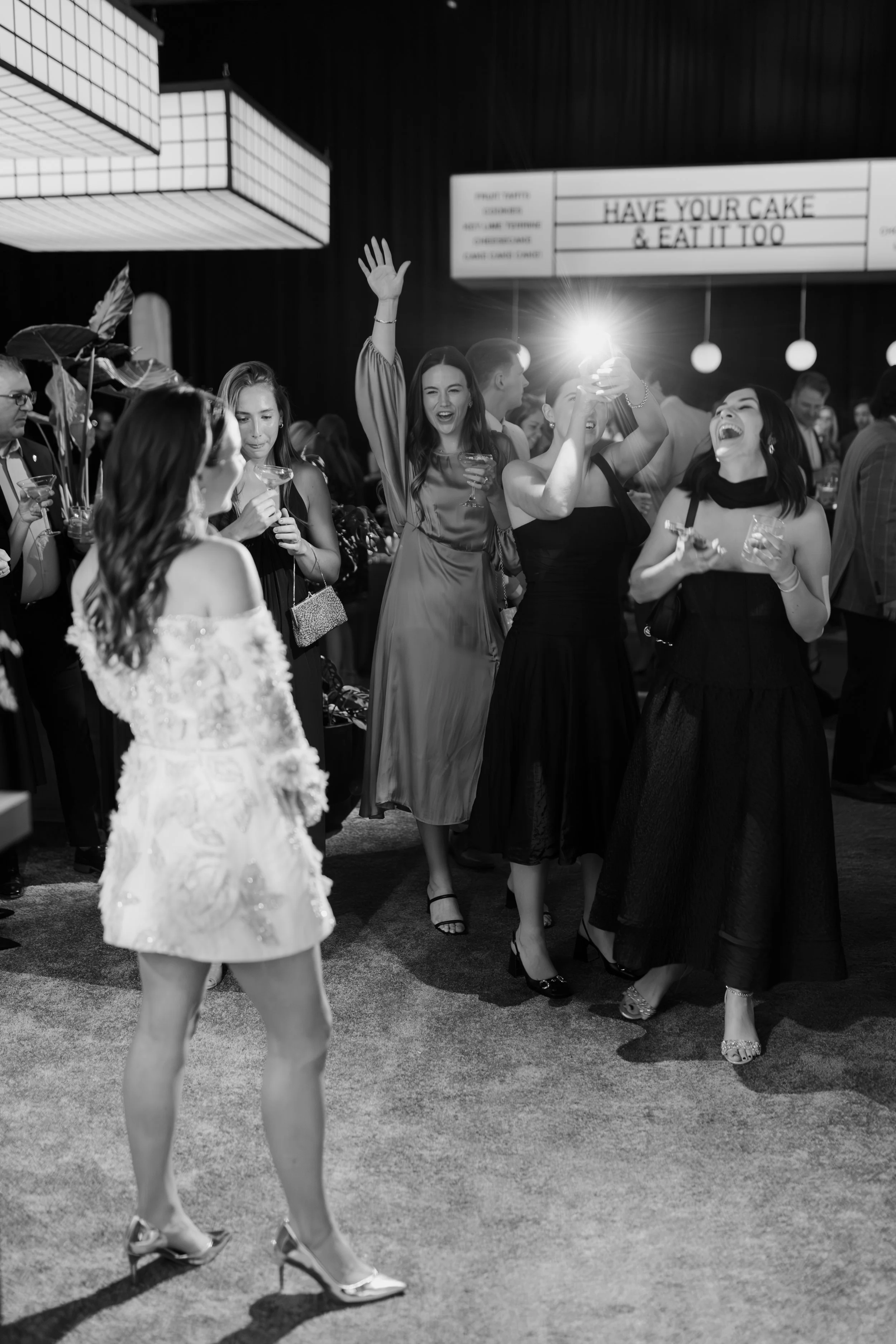 Black and white photo of a group of people at a celebration or party. In the foreground, a woman wearing a floral dress and heels is walking, while behind her, a group of women in elegant dresses are laughing, dancing, and taking selfies. There are o