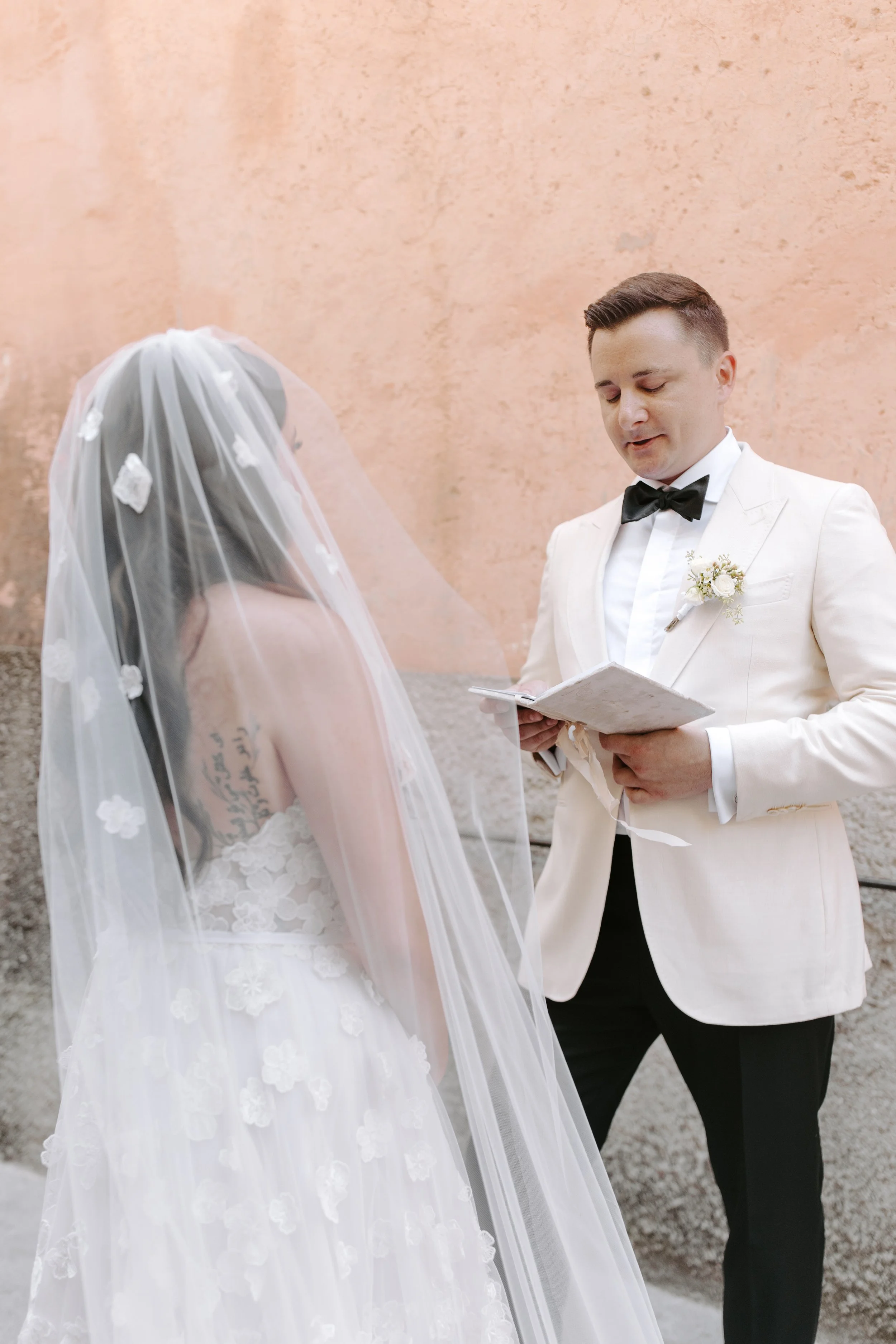 A bride and groom during their wedding ceremony, facing each other. The bride is wearing a white wedding gown with lace and a veil with floral appliqués, and has tattoos on her arm. The groom is dressed in a cream tuxedo with a black bow tie and a bo