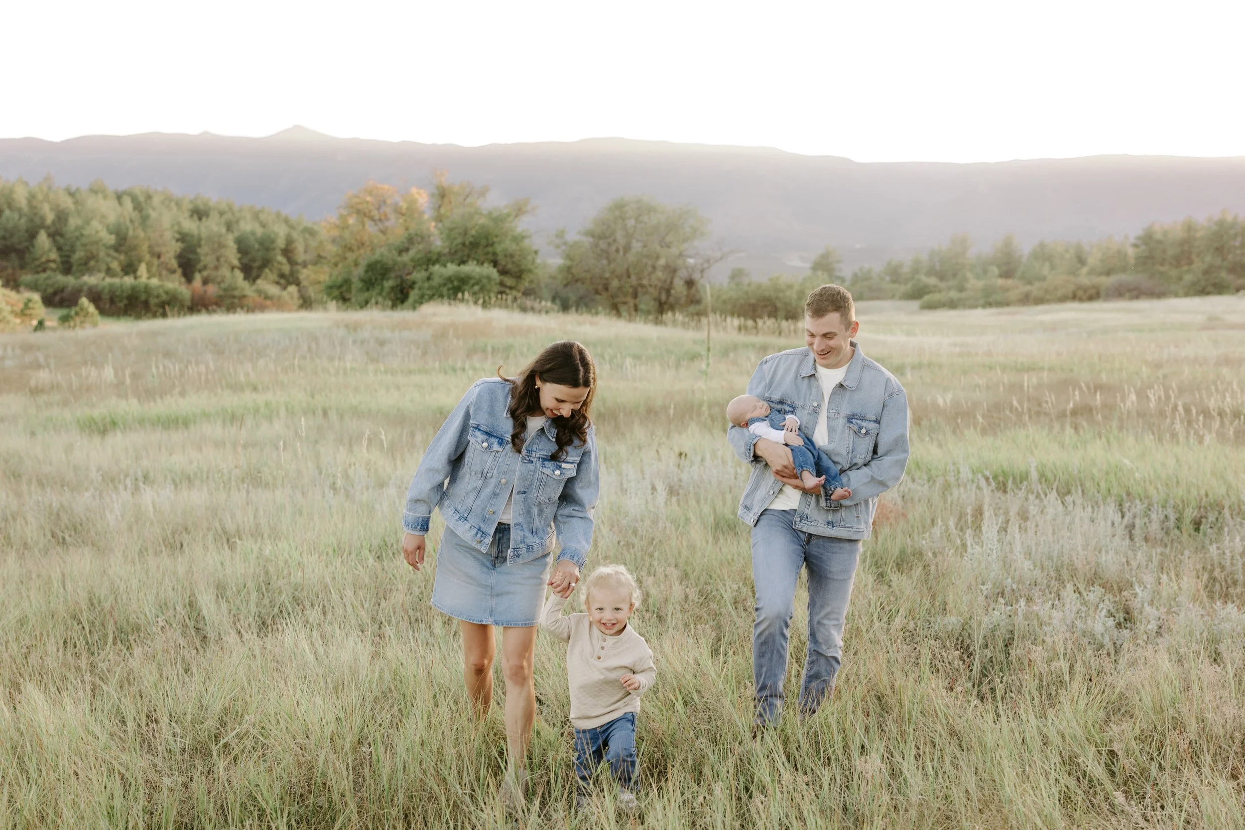 A family of four, including two children, walking through a grassy field with mountains and trees in the background during sunset.