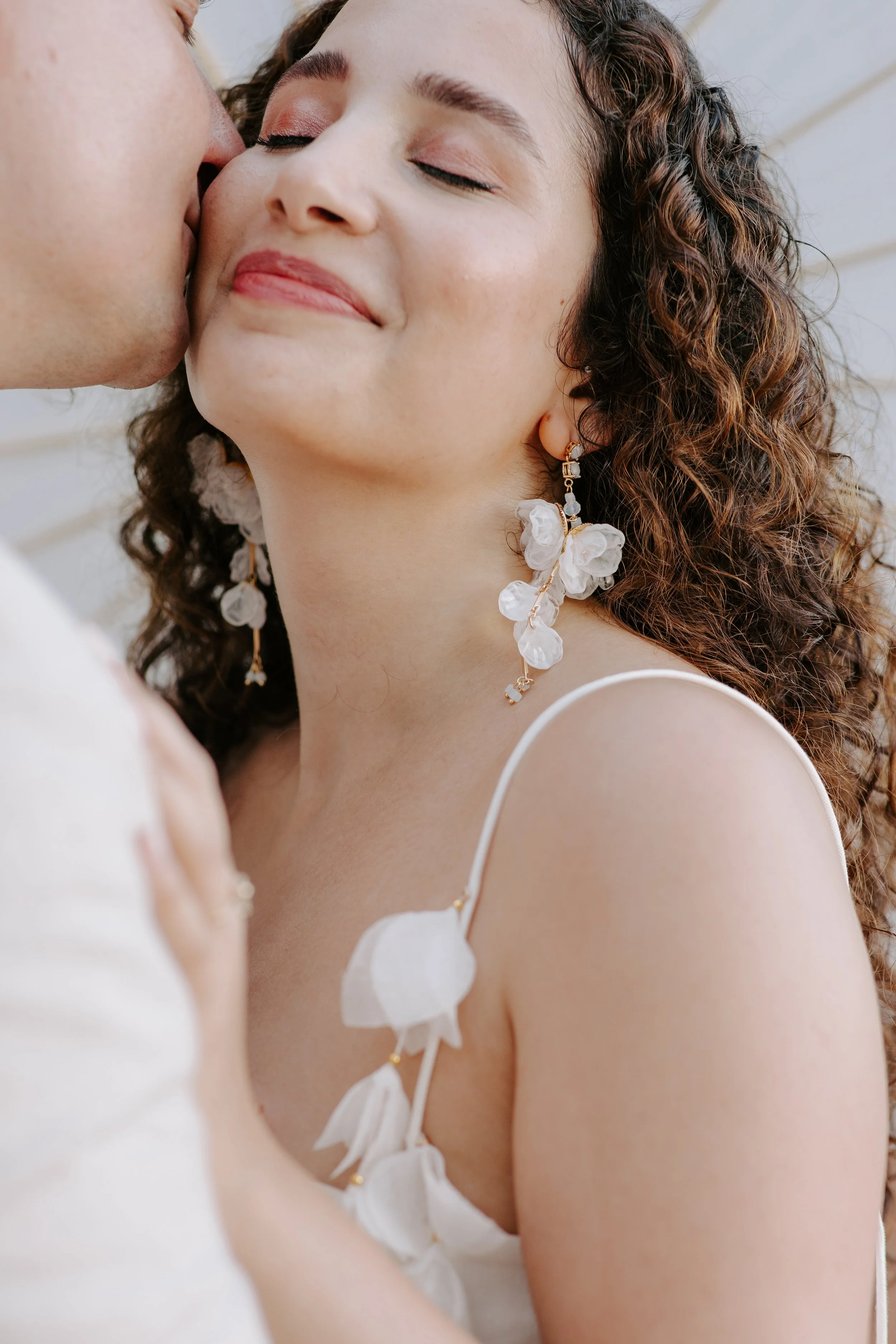 A woman with curly hair and floral earrings is smiling with her eyes closed, while someone kisses her on the cheek.