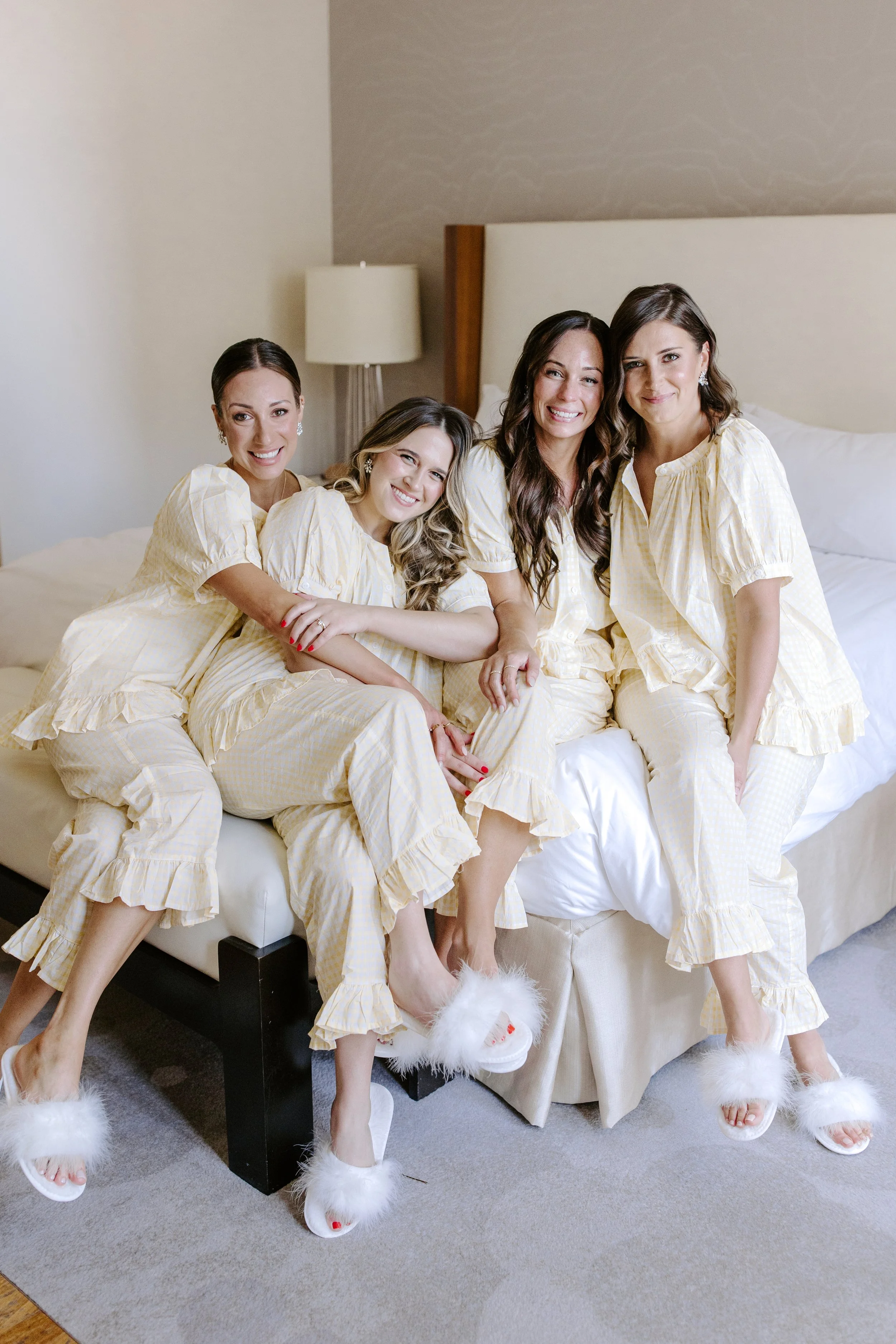 Four women sitting on a bed in matching yellow pajamas and fluffy slippers, smiling at the camera in a hotel room.
