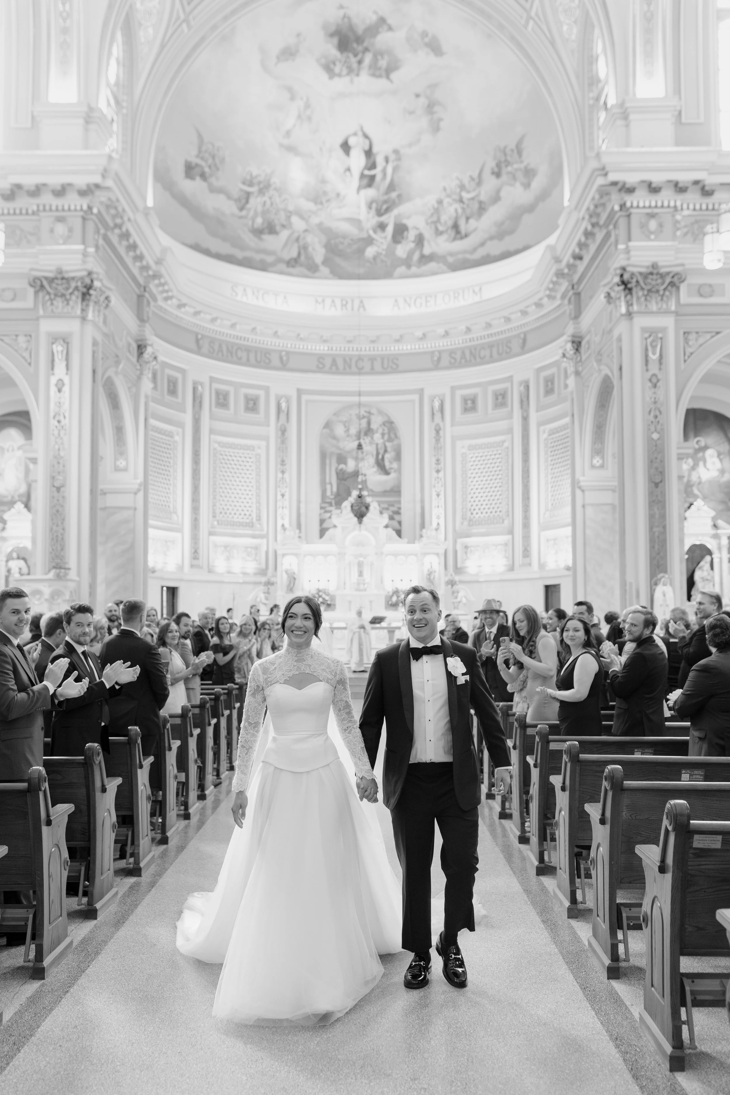 A bride and groom walking down the aisle of a church during their wedding ceremony. The bride is wearing a long white gown with lace sleeves, and the groom is dressed in a tuxedo with a bow tie. Guests are clapping and smiling on either side of the a