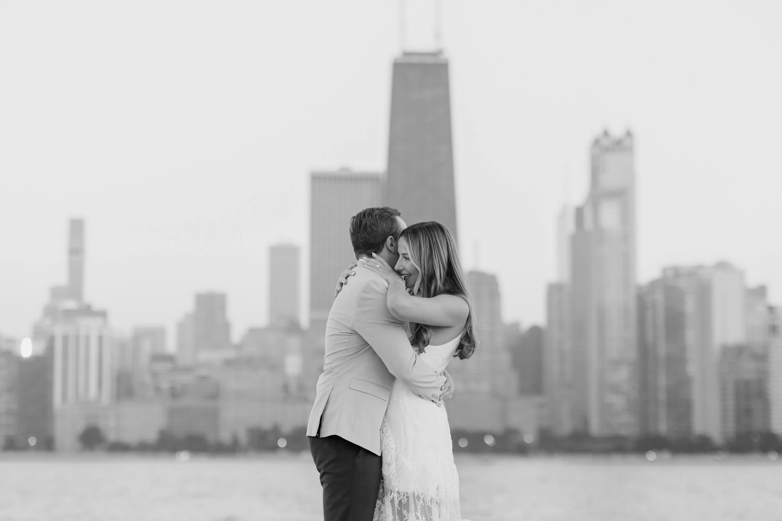 A couple hugging each other outdoors with a city skyline in the background, viewed in black and white.