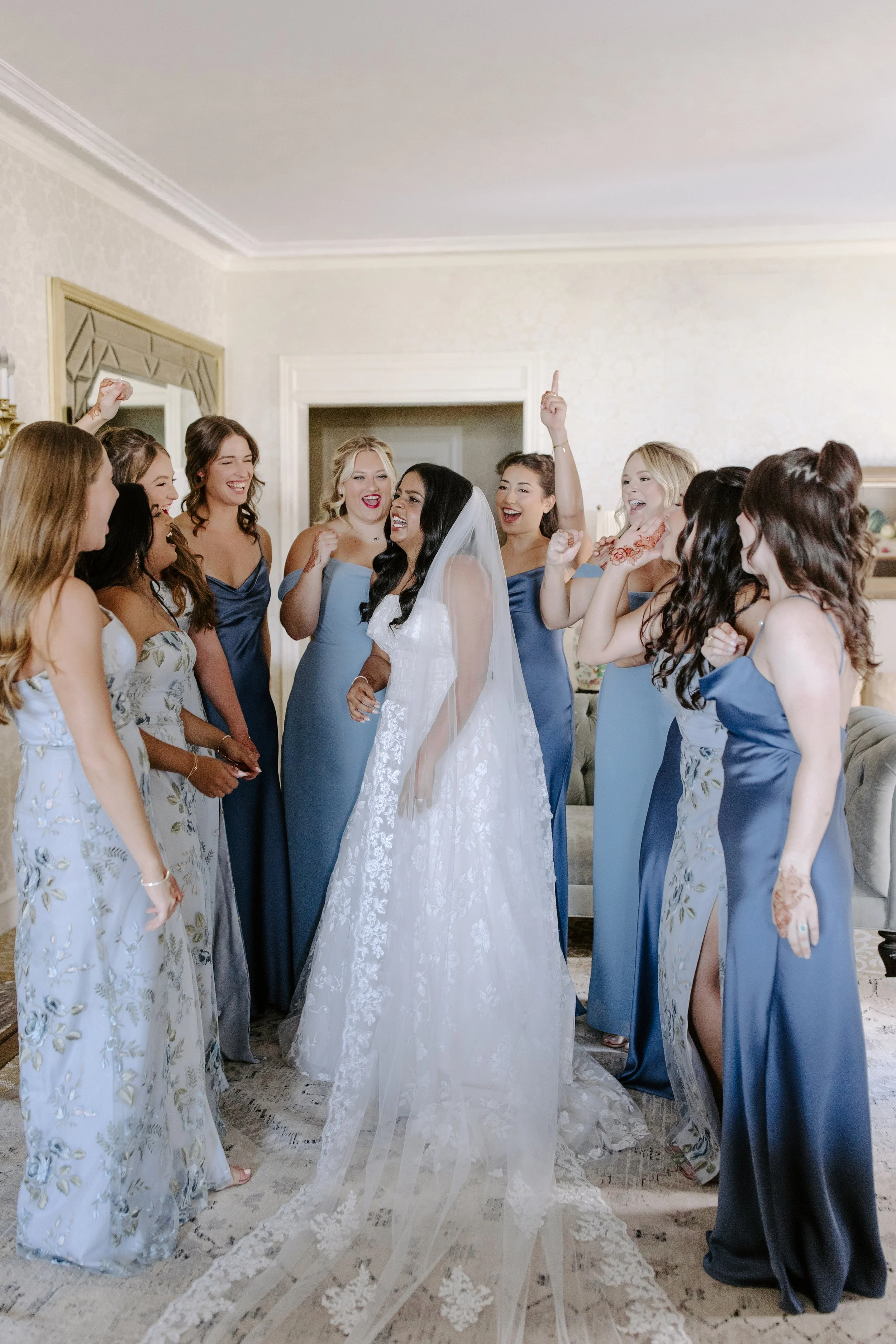 A bride in a white wedding gown and veil is surrounded by a group of women in matching blue and floral bridesmaid dresses inside a decorated room.