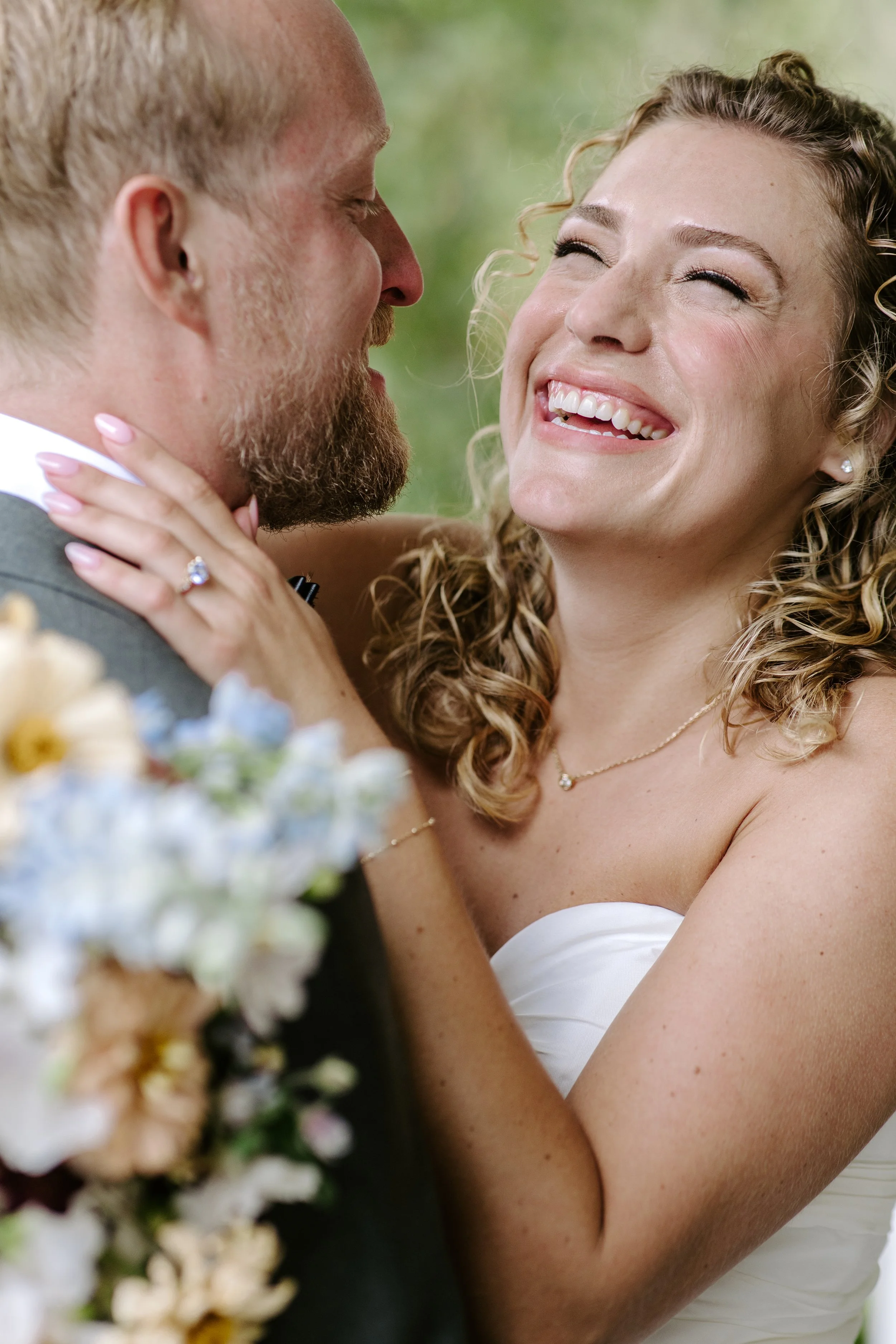 A smiling woman with curly hair looking at a man with a beard. They are close together, possibly on their wedding day.